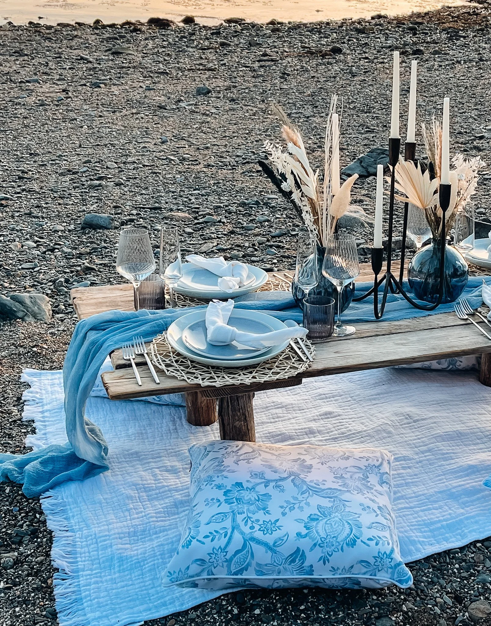 Beach picnic setup with a low wooden table, plates, glasses, cutlery, decorative vases with dried flowers, black candle holders with white candles, a blue cloth, a white quilt, and a blue floral pillow on rocky ground near water.