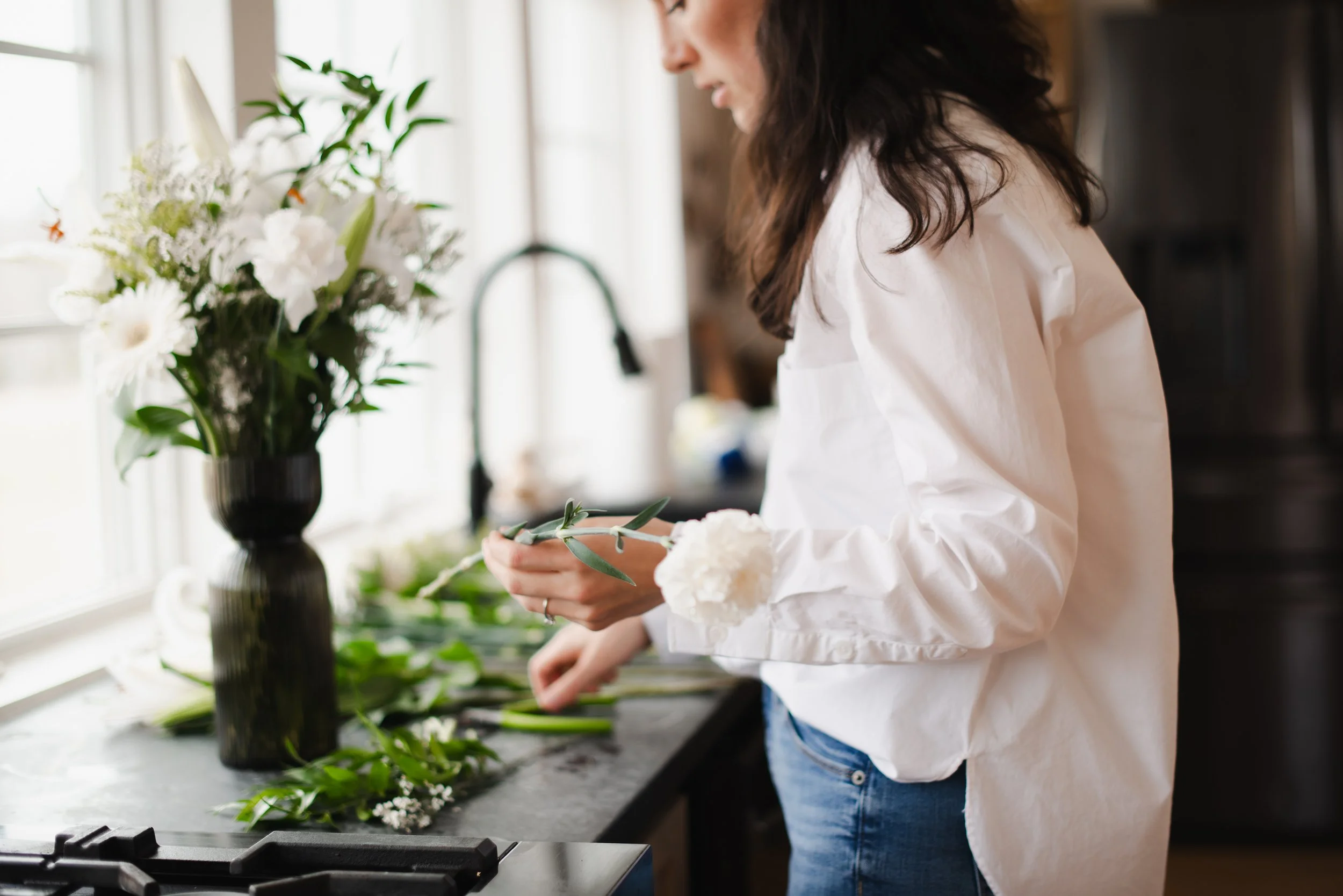 A woman in a white shirt arranging white flowers and greenery on a kitchen counter with a black vase and a large flower arrangement.
