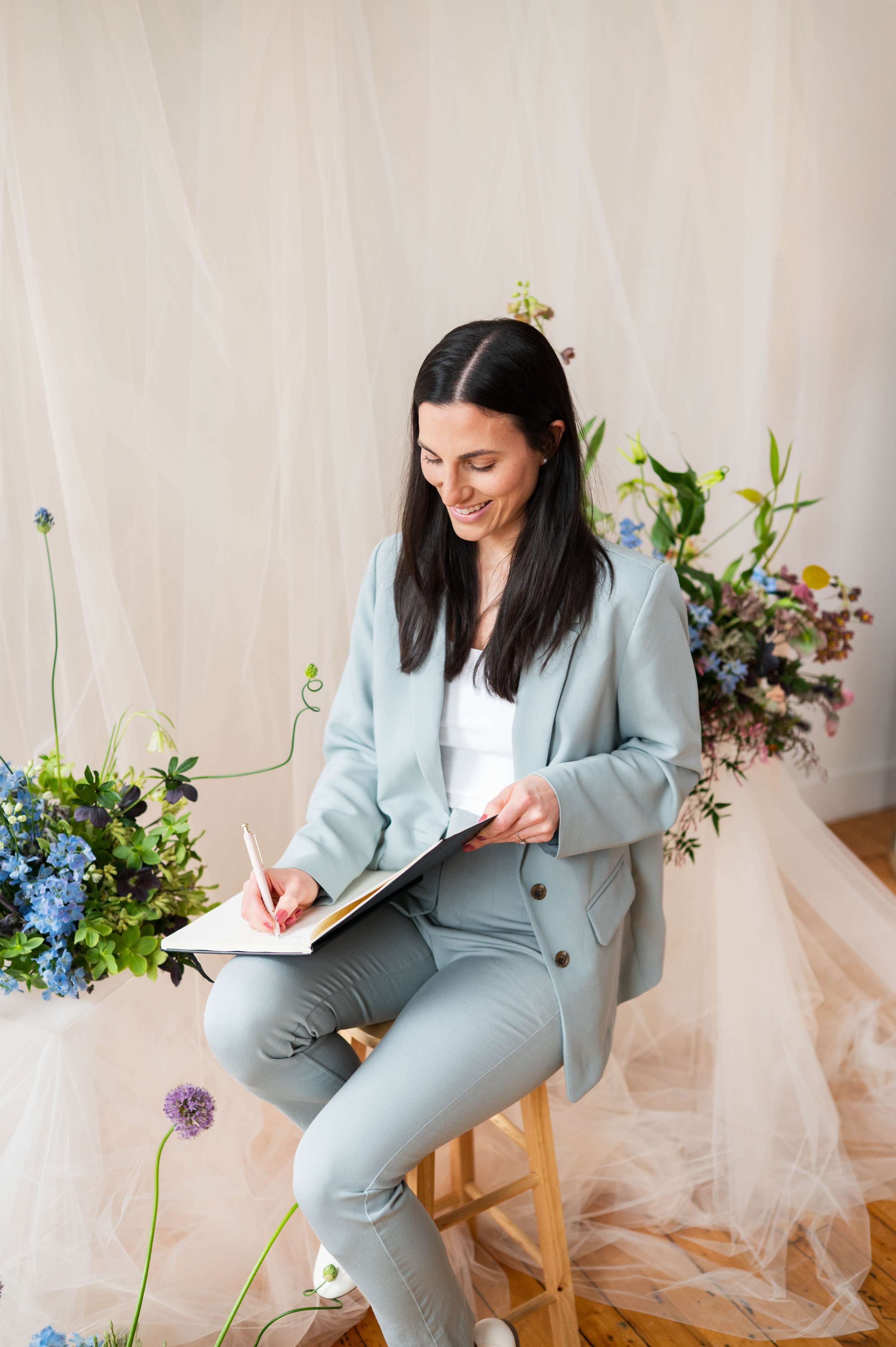A woman with long dark hair, dressed in a light gray suit, sitting on a wooden stool, smiling, and writing notes in a notebook. She is surrounded by floral arrangements with colorful flowers and greenery, with light-colored curtains and fabric draped in the background.