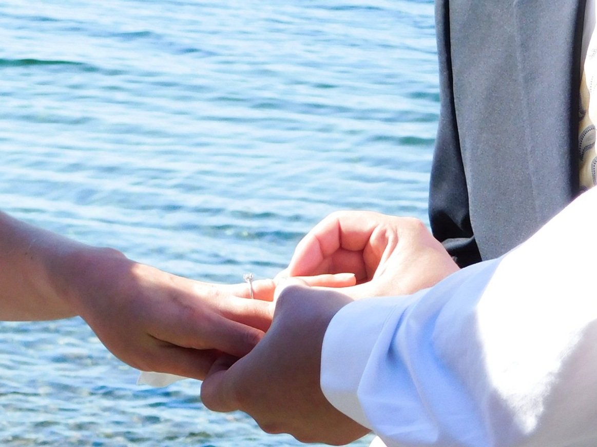 Close-up of a couple holding hands during a wedding ceremony by the water, with the bride wearing a ring and the groom in a suit.