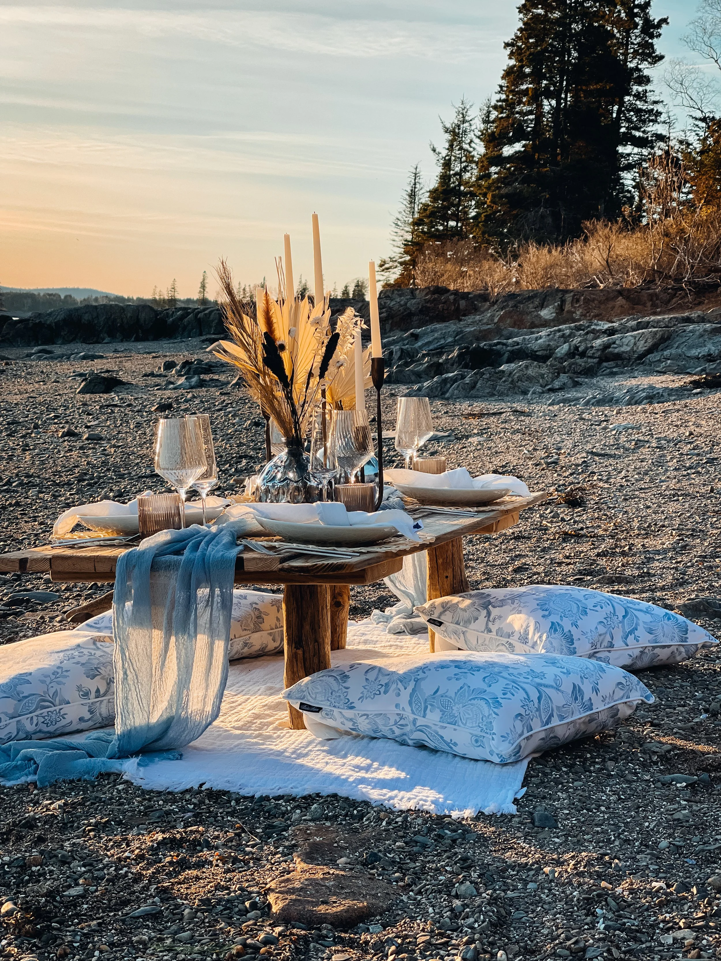 A beach picnic setup with a low wooden table, pillows on the sand, and a white blanket, set with wine glasses, plates, candles, and a floral centerpiece, during sunset with trees in the background.