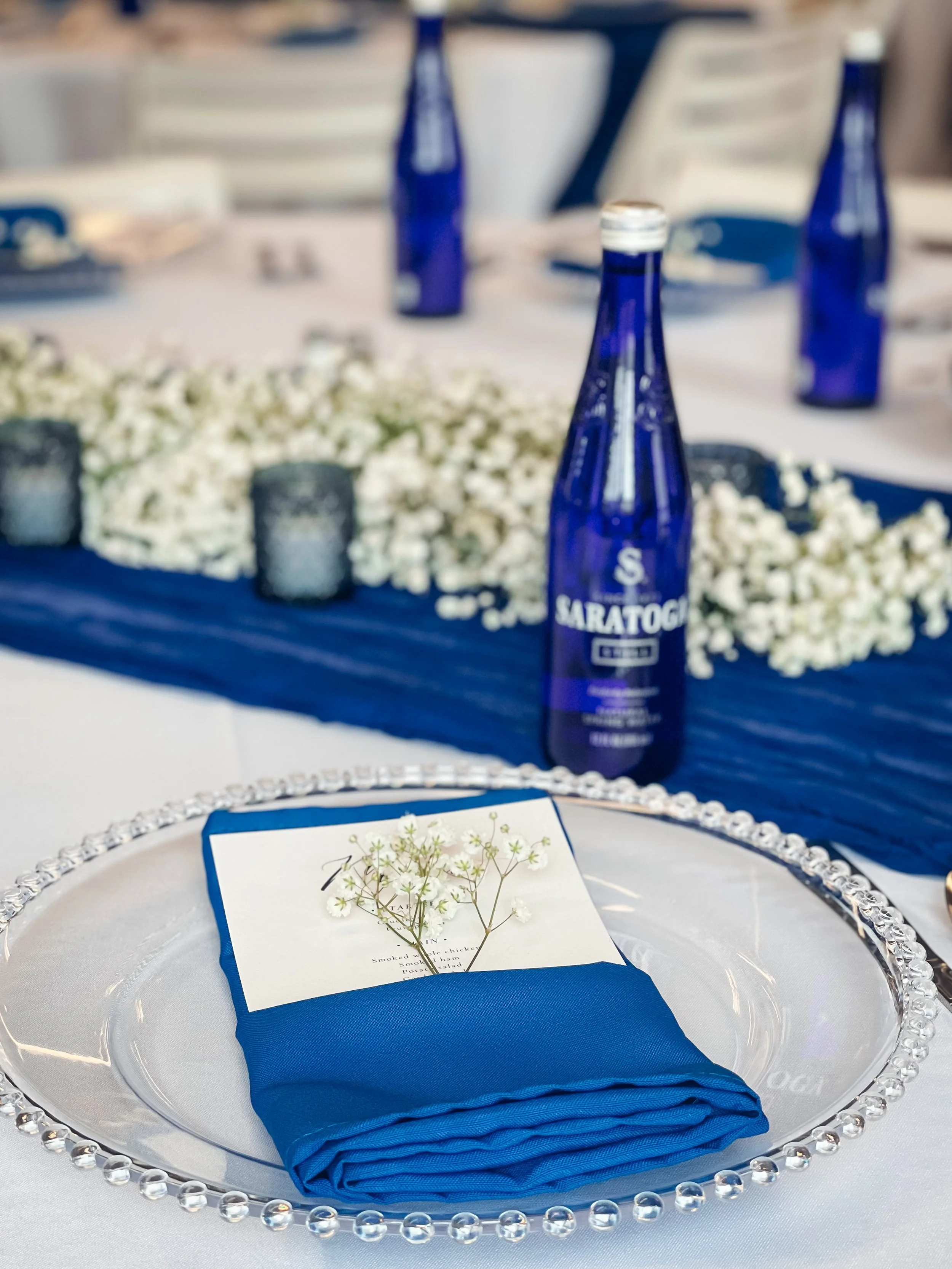 Table setting with a glass charger, a folded blue napkin, a menu card tucked under the napkin, and small white flowers on top. In the background, there are blue bottles labeled "Saratoga" and a floral centerpiece with white flowers.