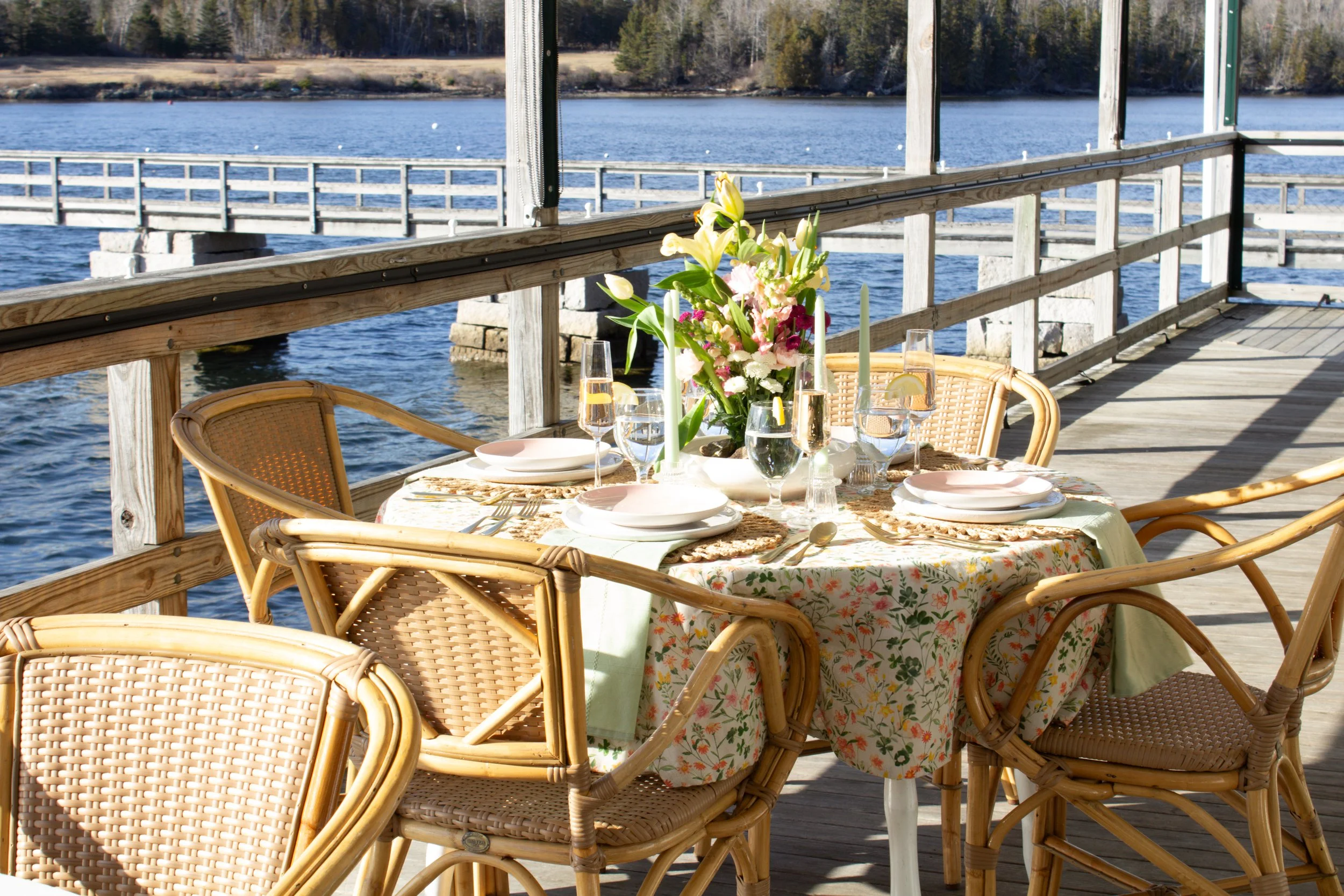 An outdoor dining table set with a floral tablecloth, plates, glasses, cutlery, and a central floral arrangement, situated on a wooden pier overlooking a body of water in daylight.