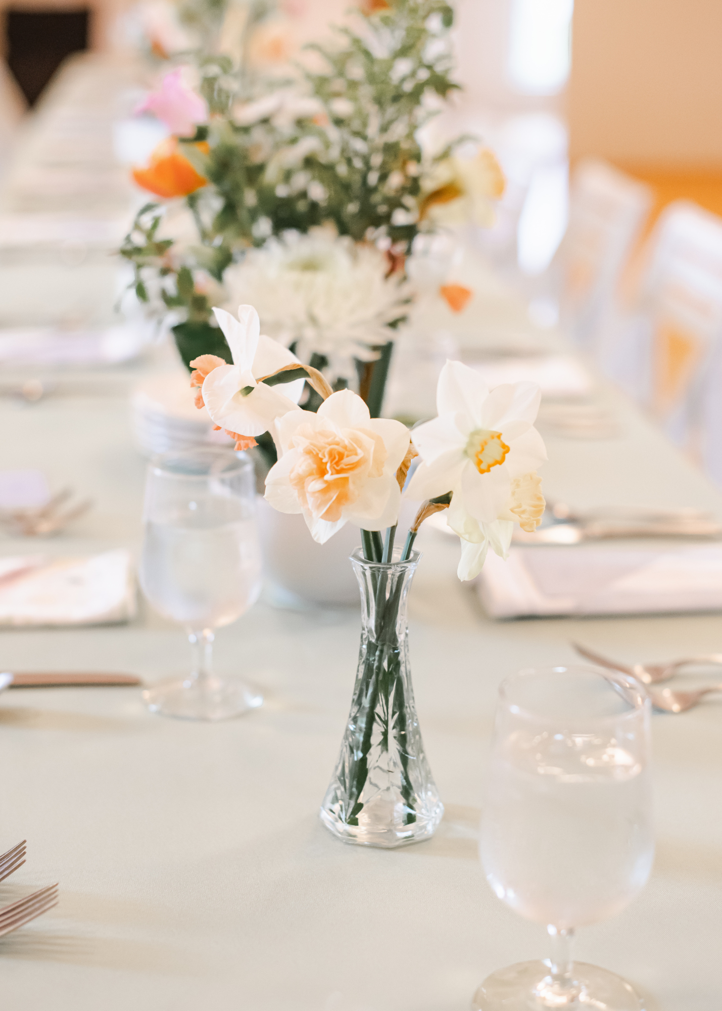 Elegant table centerpiece with white and peach flowers in a clear glass vase, set on a dining table with water glasses and silverware, in a well-lit room.