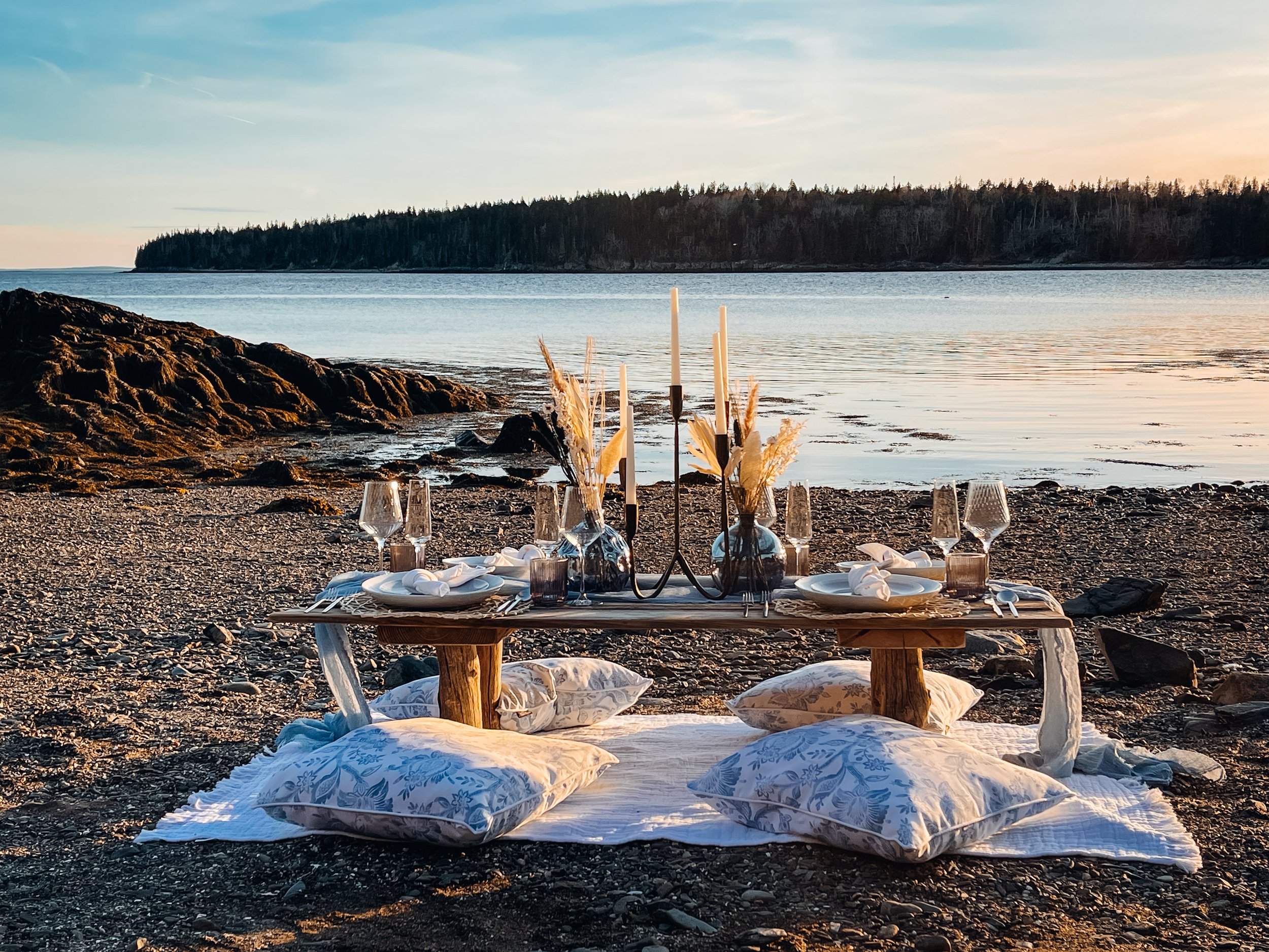 Beachside outdoor dining setup with a low wooden table, candles, glassware, and pillows on a white cloth, overlooking calm water and a treeline in the distance at sunset.