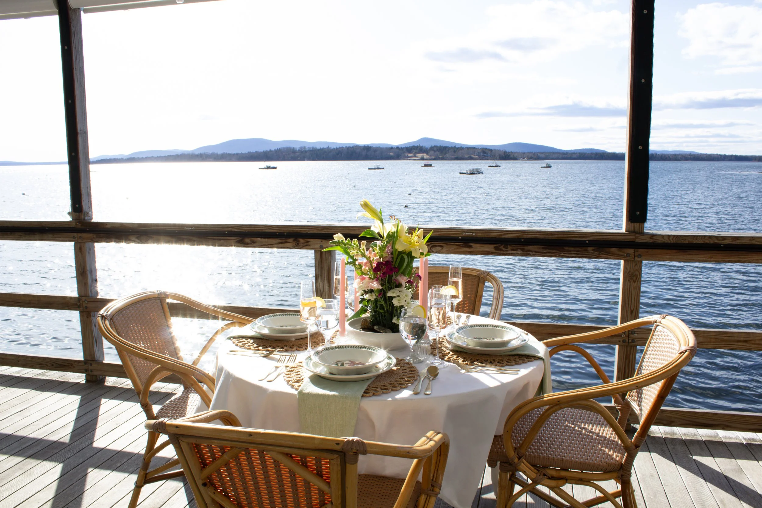 A round dining table set for four on a balcony overlooking a lake with mountains in the distance. The table has a white tablecloth, floral centerpiece, plates, glasses with lemon slices, and cutlery. Four wicker chairs surround the table, and the scene is lit by natural sunlight.