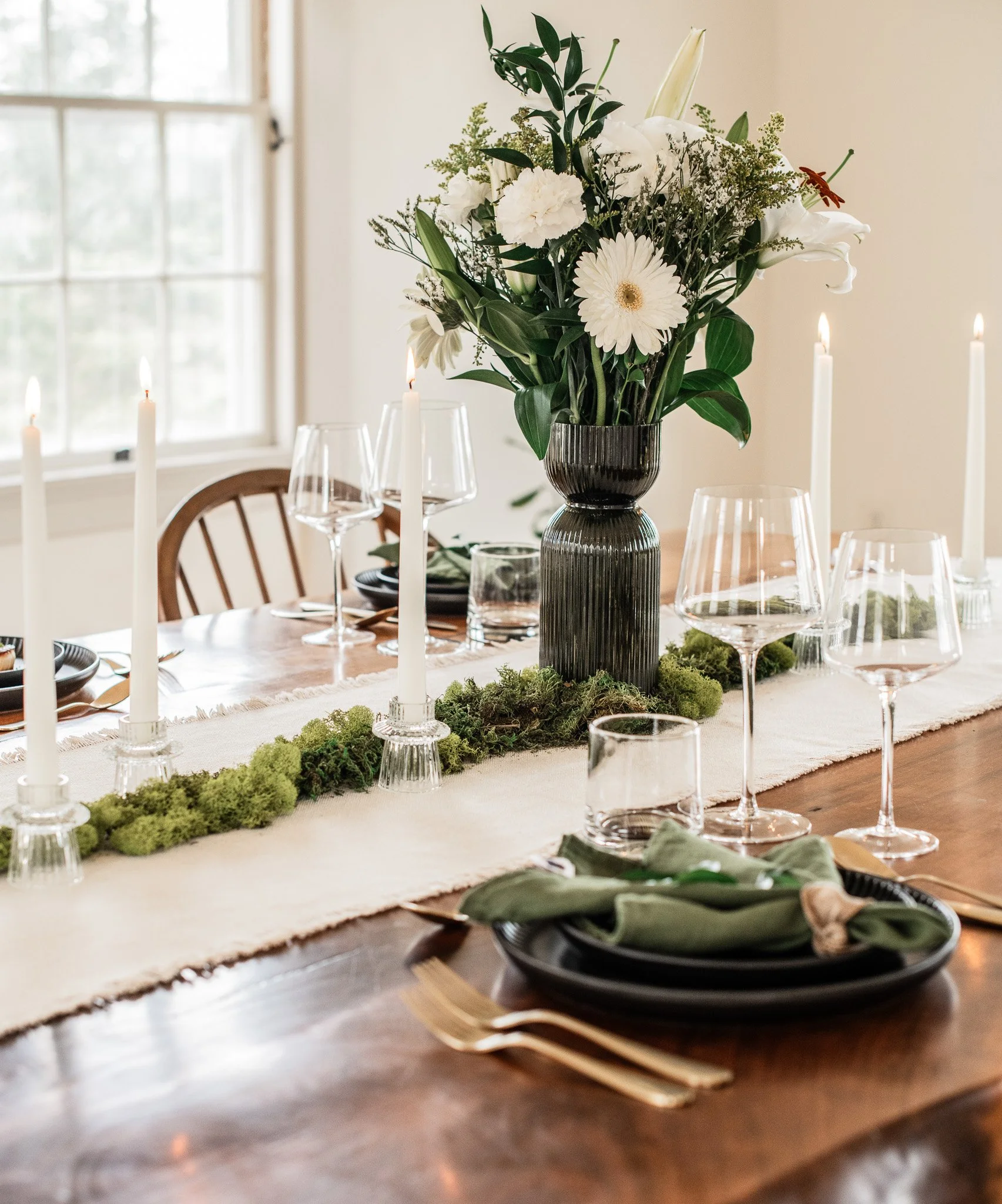 Close-up of a decorated dining table with a large floral arrangement, candles, green moss, and elegant place settings.