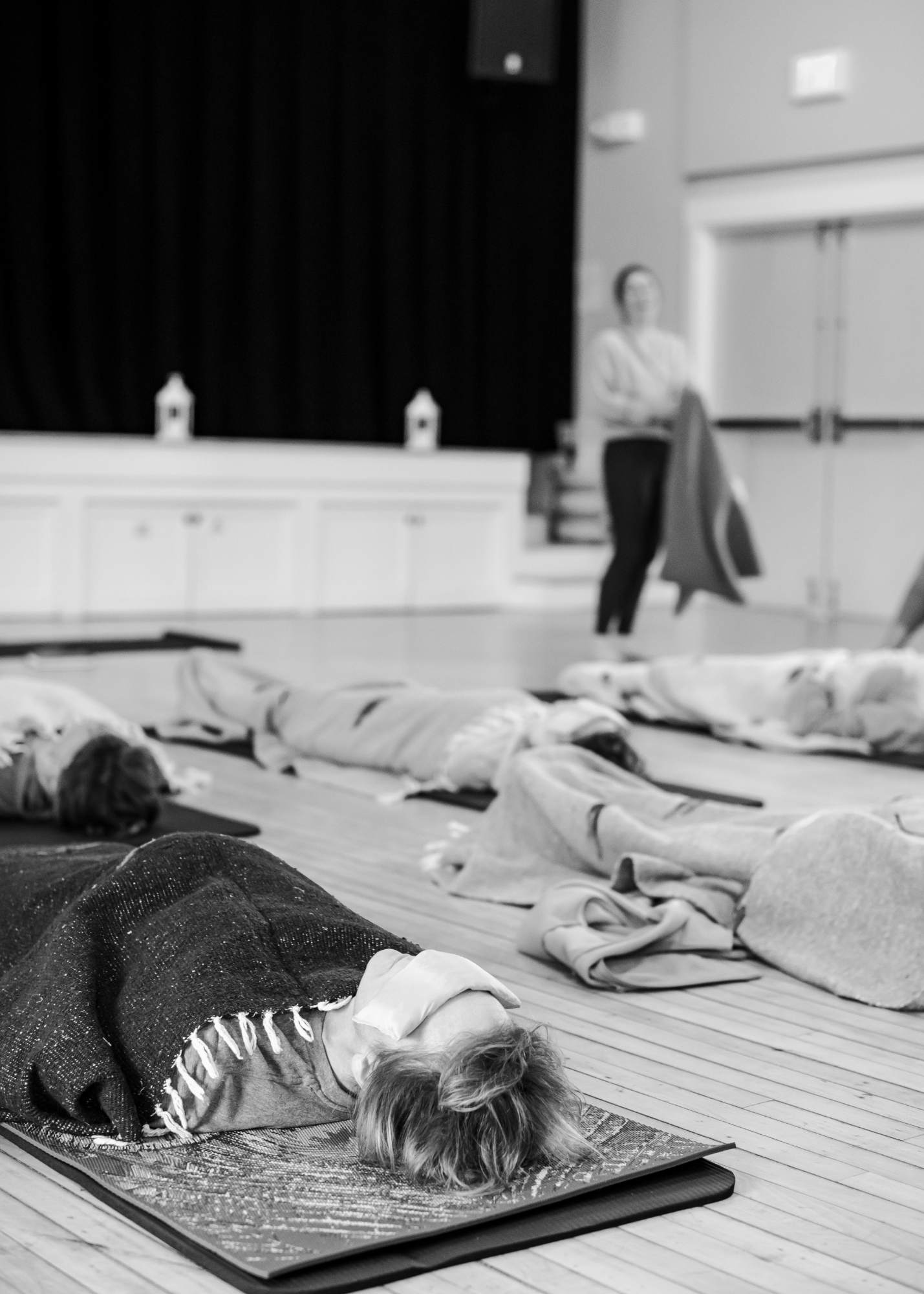 A person lying on a yoga mat with their eyes closed, wearing a face mask, in a room with folding mats and another person standing in the background.