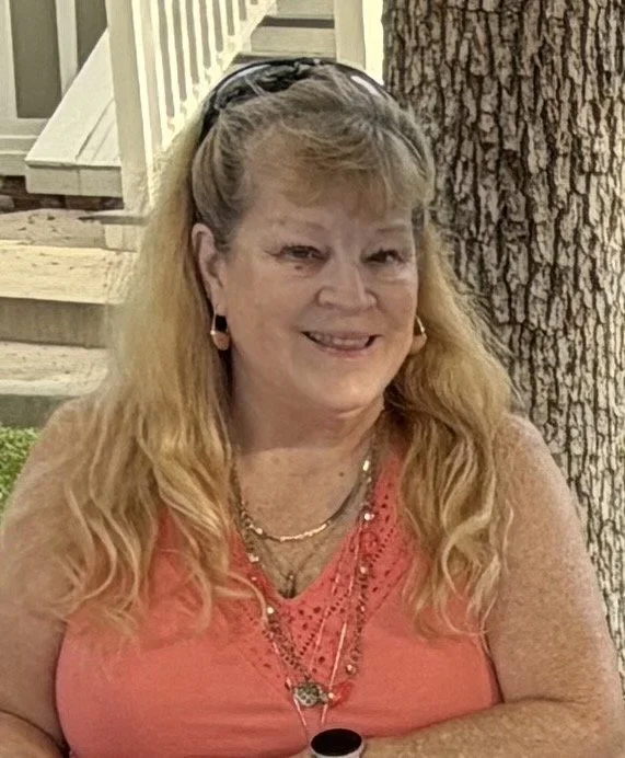 A woman with long, light brown hair and fair skin, wearing a coral-colored top with lace detail, gold hoop earrings, and layered necklaces, standing outdoors near a tree with a porch and stairs in the background, smiling at the camera.
