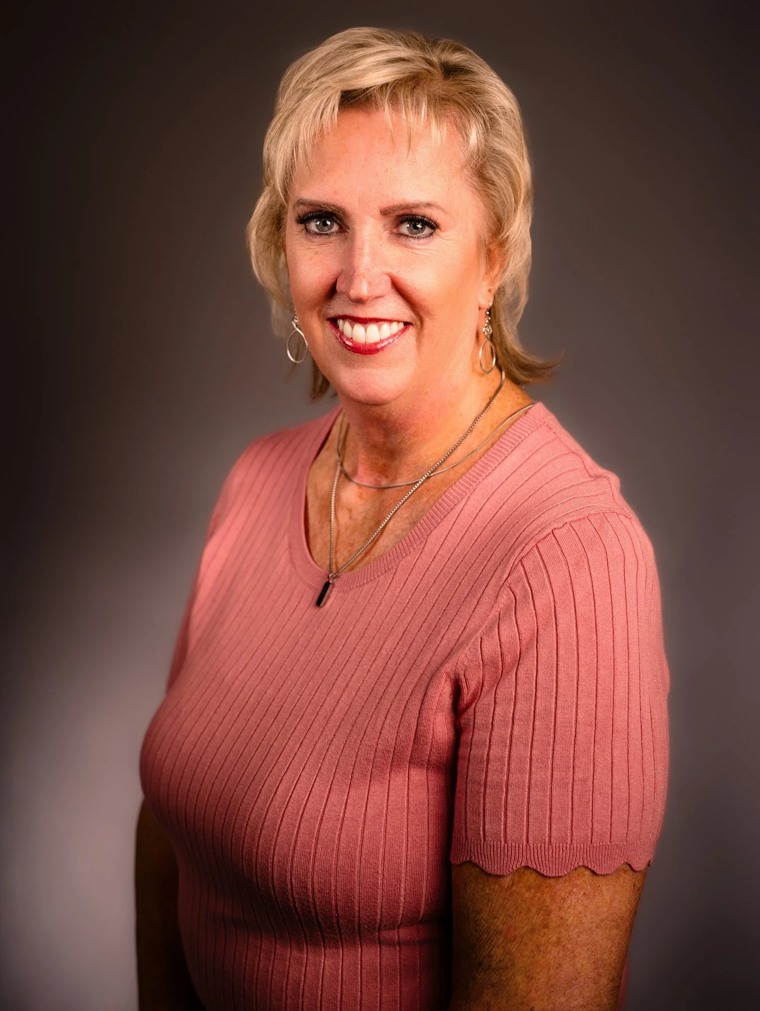 A woman with short blonde hair, wearing a pink ribbed top with scalloped sleeves, and jewelry including earrings and necklaces, smiling at the camera against a dark gradient background.