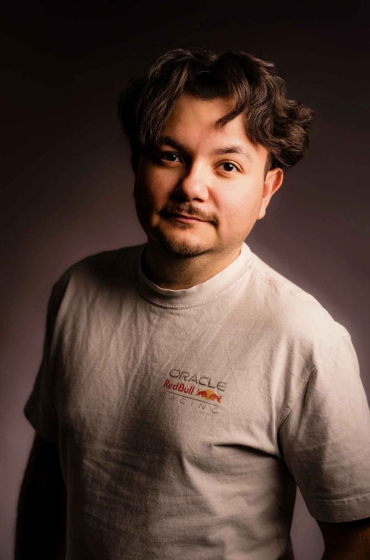 A young man with dark, tousled hair and a trimmed beard wearing a beige t-shirt with the Oracle Red Bull Racing logo, standing against a dark gradient background.