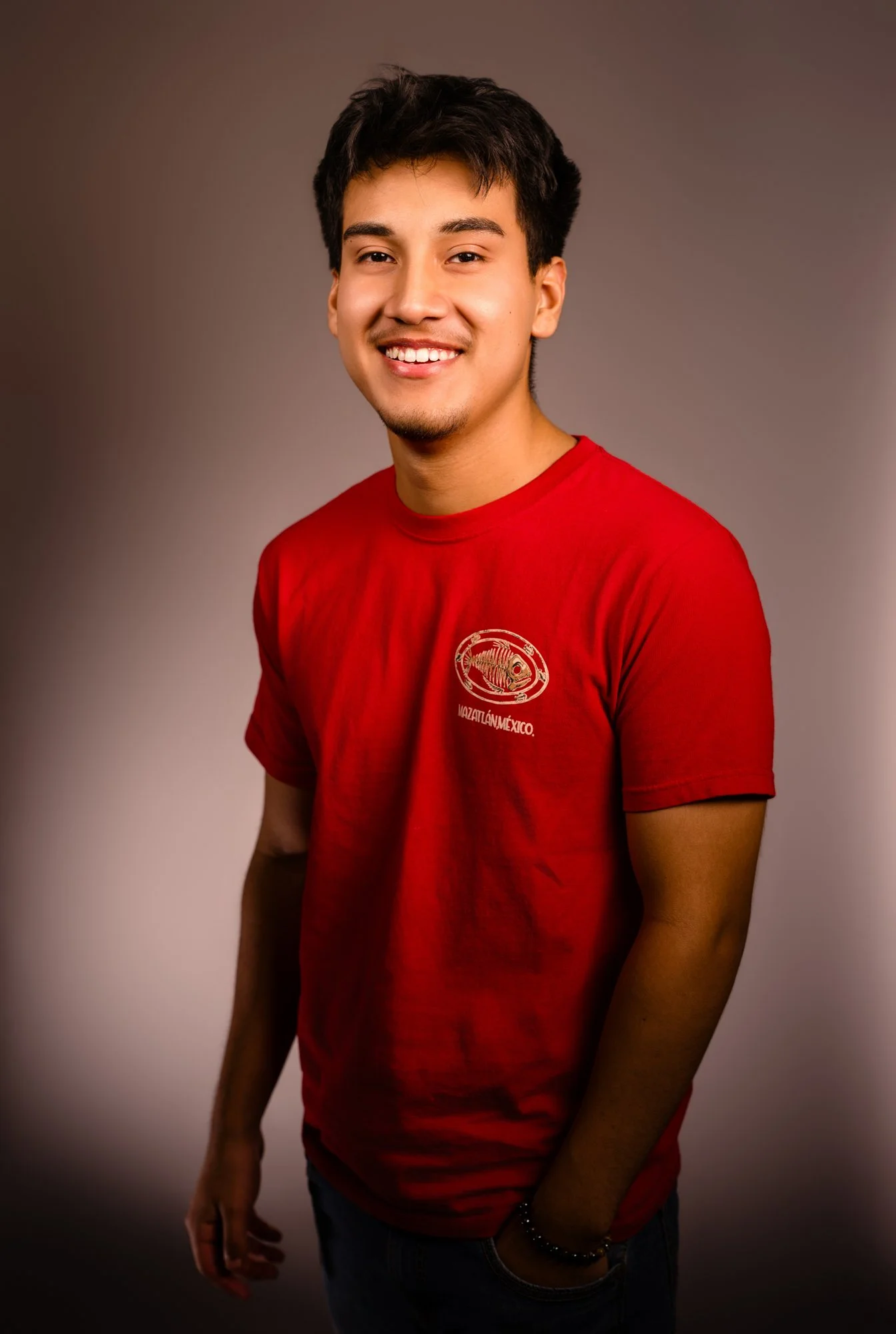 A young man with short black hair, smiling, wearing a red T-shirt with a logo and text, and standing against a plain background.