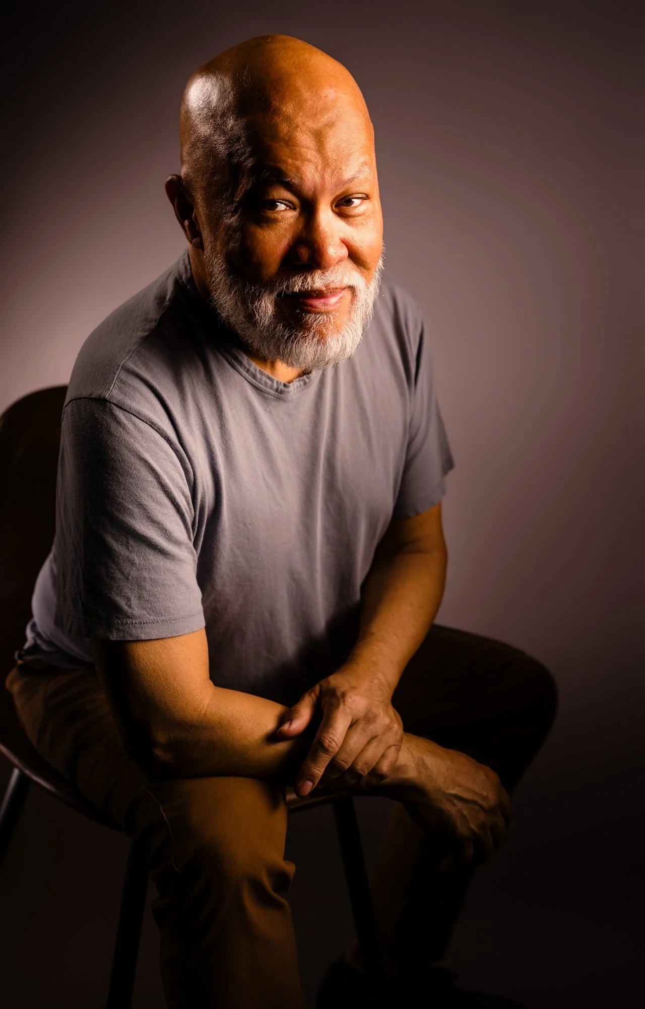 Portrait of an older man with a bald head and gray beard, wearing a gray t-shirt, sitting on a chair against a dark gradient background.
