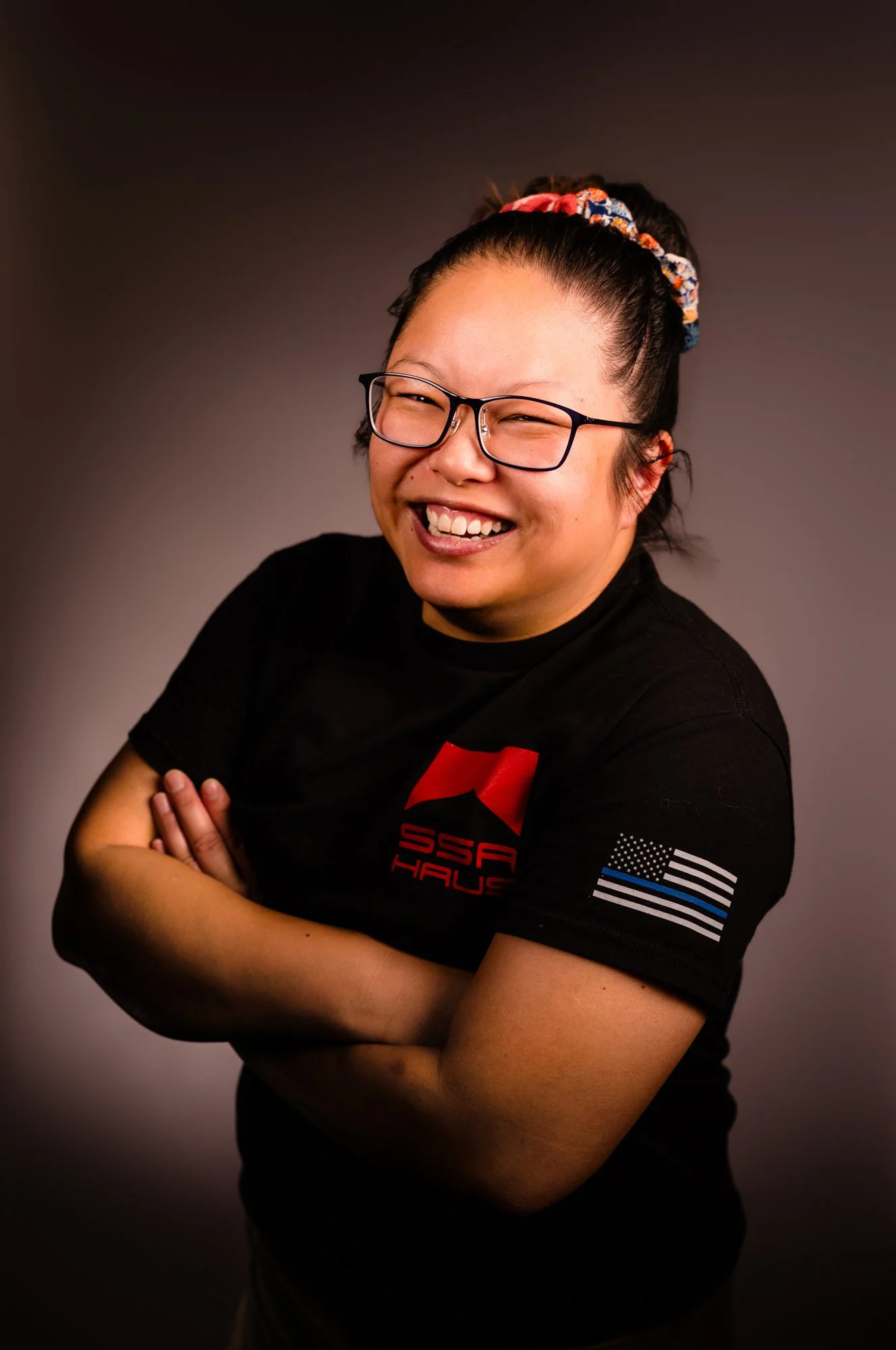 Woman with glasses smiling, arms crossed, wearing a black T-shirt with red, white, and blue US flag and American flag patches on the sleeves, against a dark studio background.
