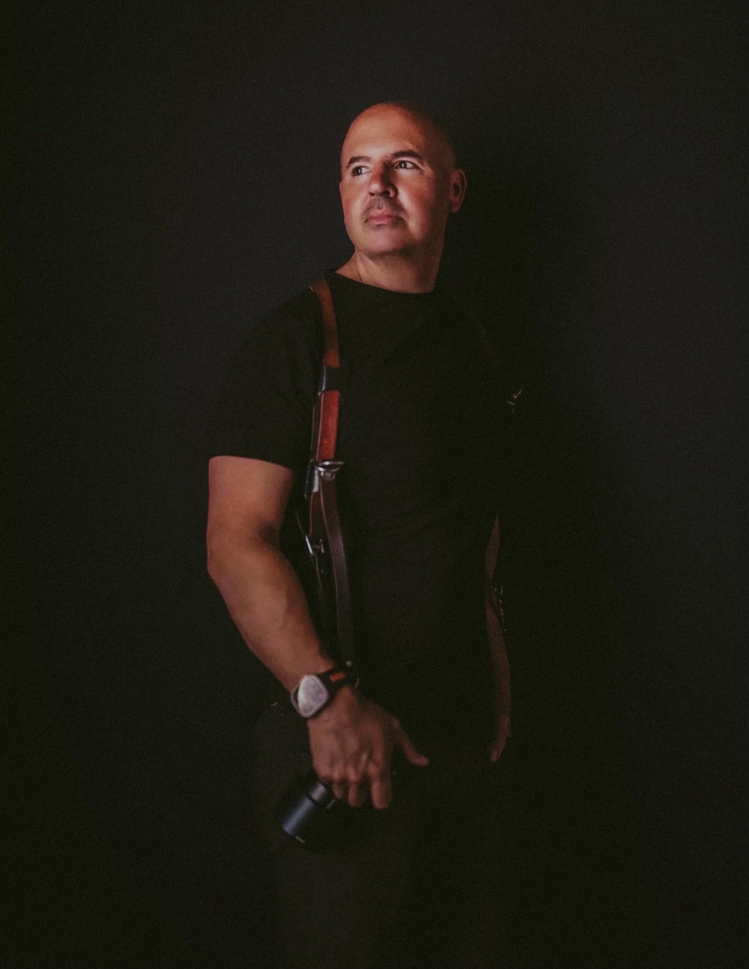 A man wearing a black T-shirt, a watch, and a backpack, holding a trekking or hiking stick, standing against a dark background.