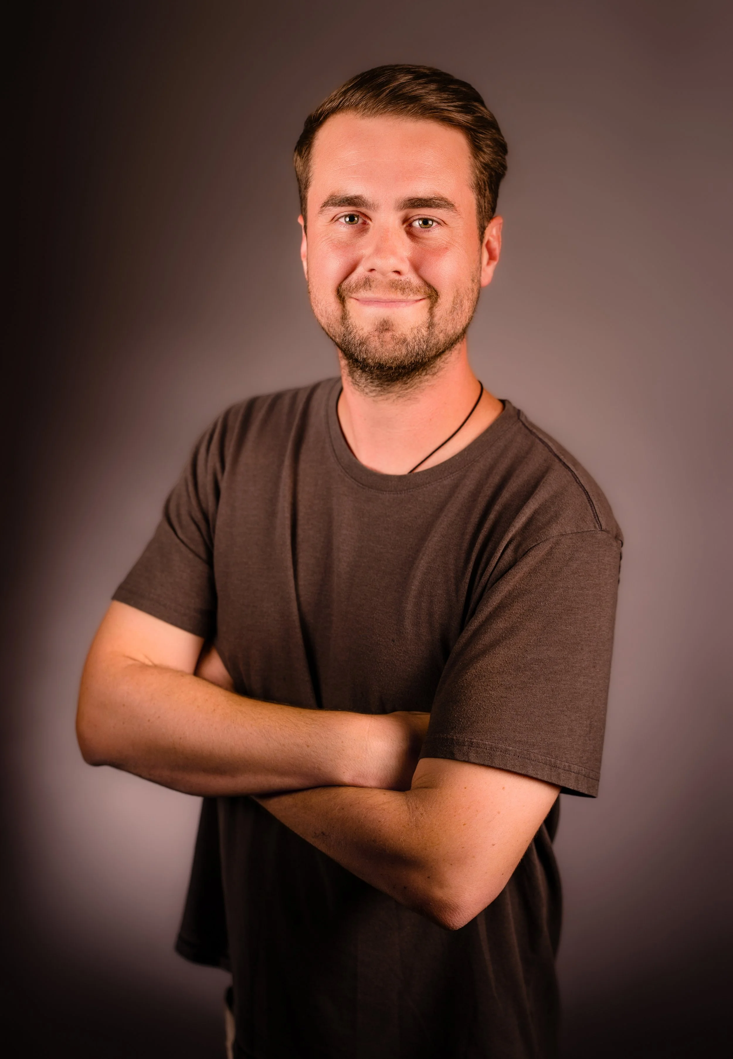 A young man with short brown hair, light skin, and a beard standing with arms crossed, smiling slightly, wearing a brown t-shirt and a black necklace, against a plain dark background.