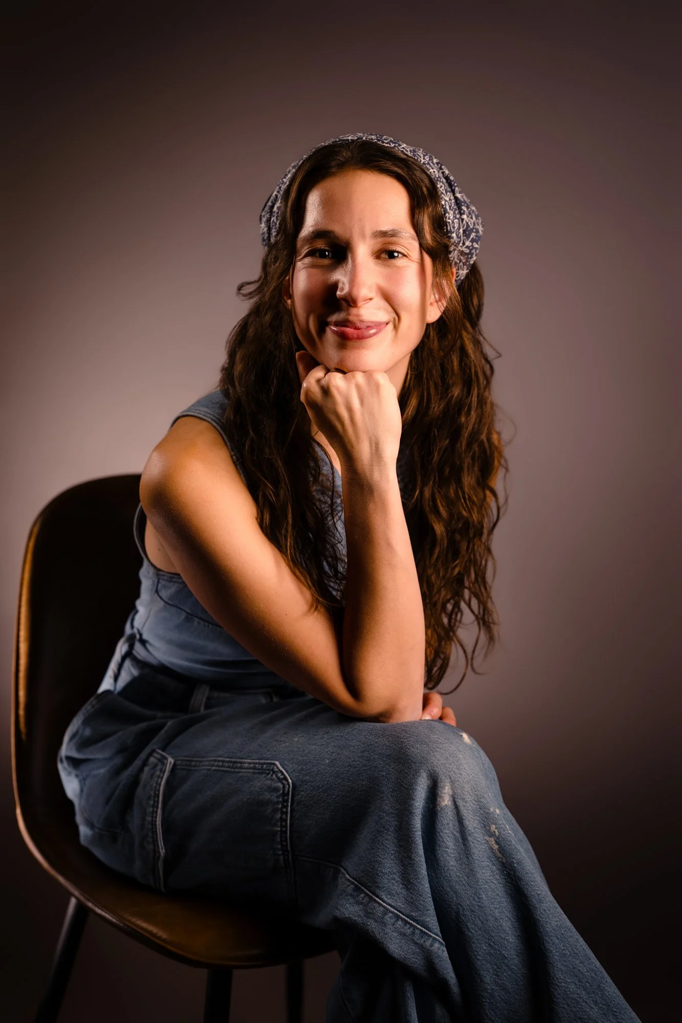 Young woman with long curly hair, wearing a denim sleeveless top and a headscarf, sitting on a black chair against a dark background, smiling and resting her chin on her hand.