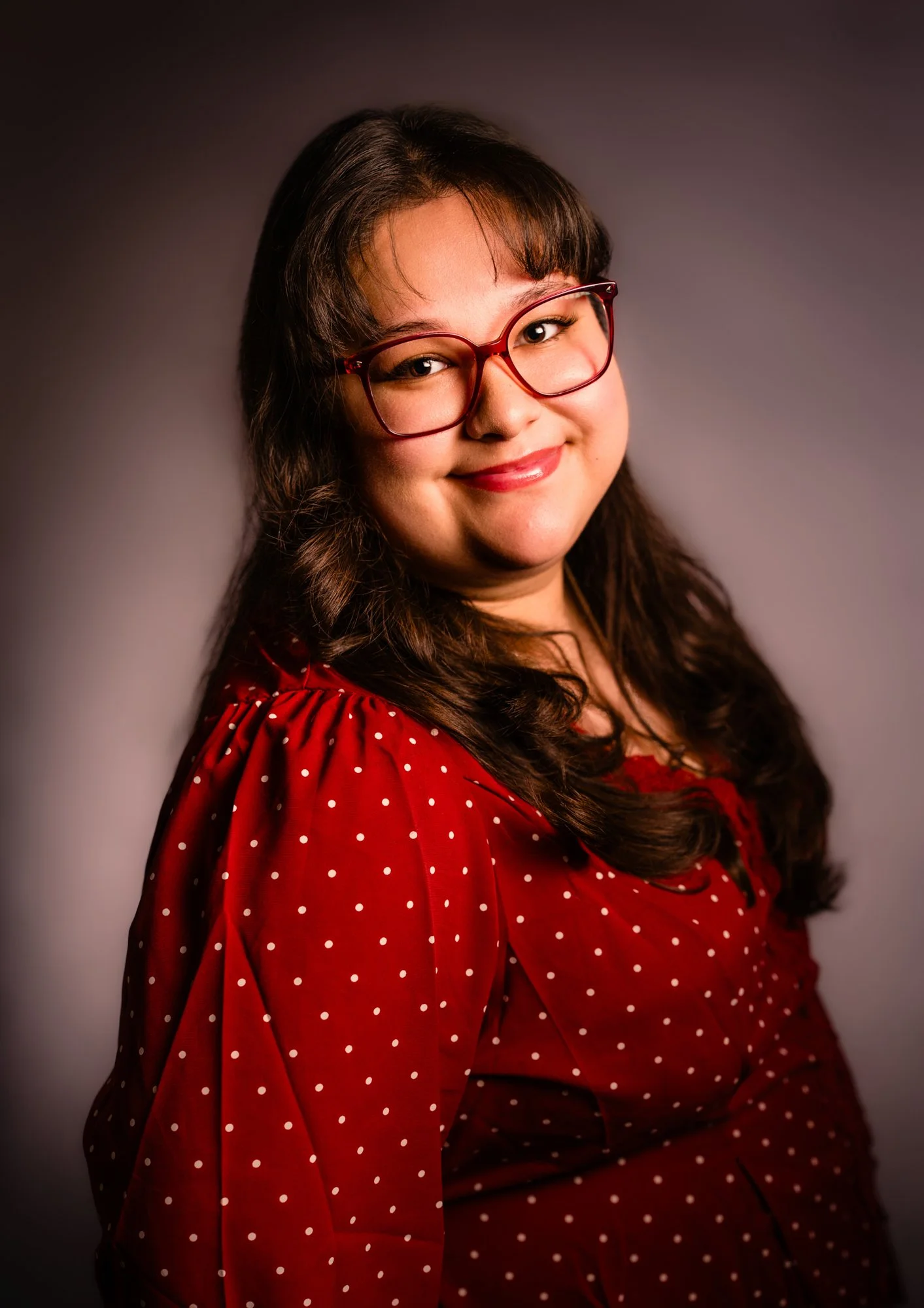 A woman with dark wavy hair and red glasses smiles at the camera, wearing a red polka-dot blouse against a dark gradient background.