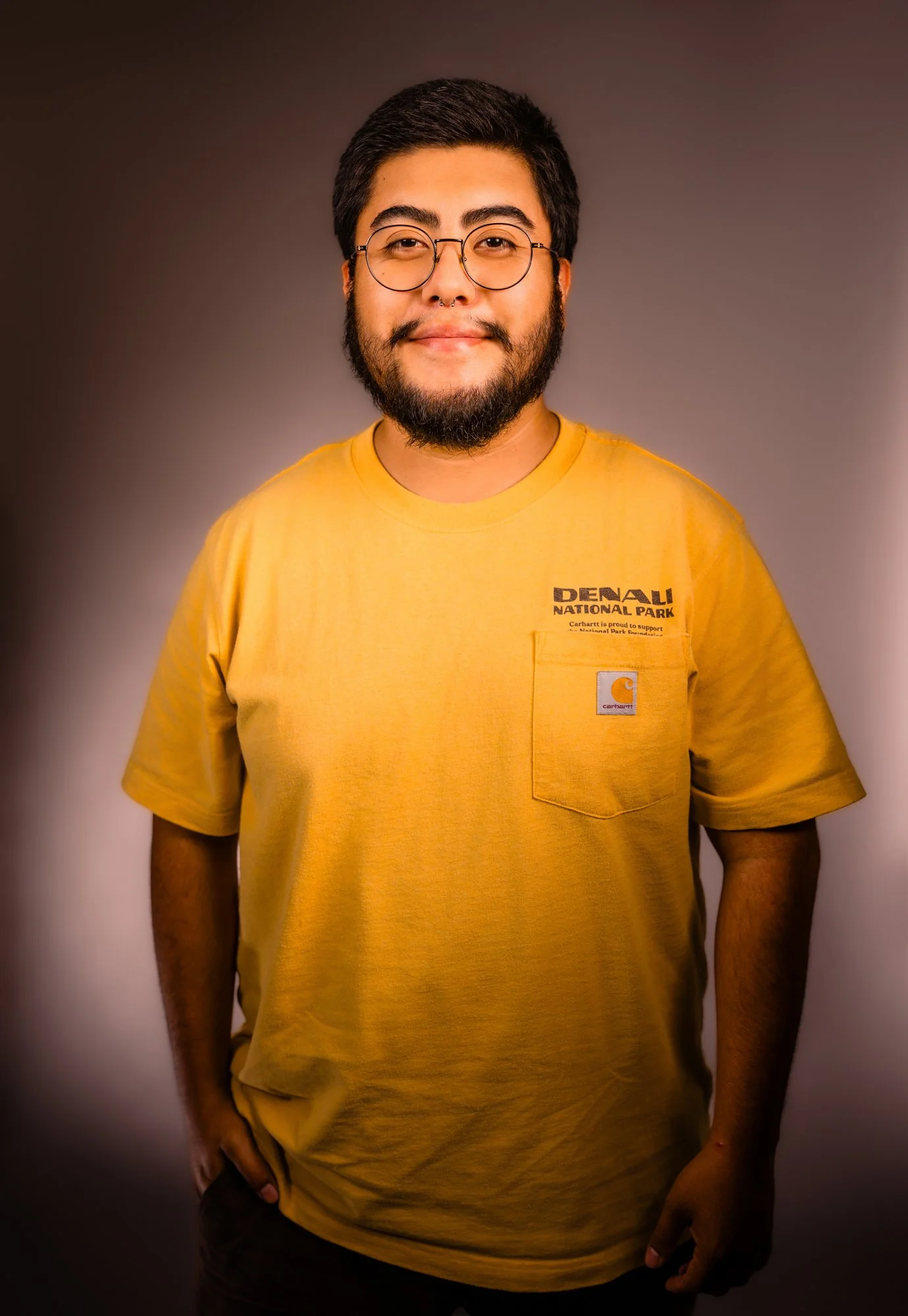 Portrait of a young man with dark hair, beard, glasses, and a septum piercing, wearing a yellow T-shirt with a pocket that has a Carhartt logo, standing against a brown and dark gradient background.