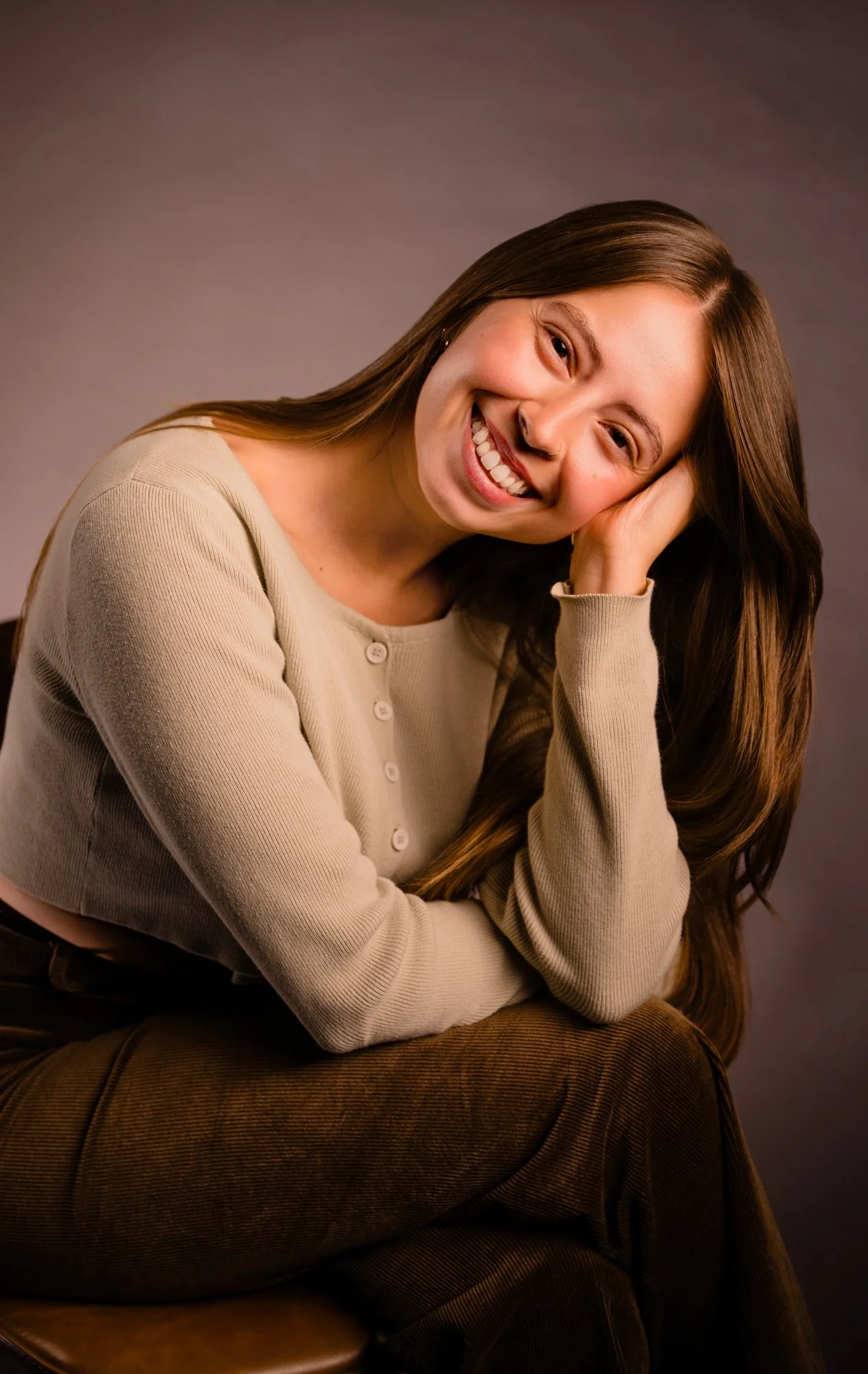 A young woman with long brown hair smiling, resting her head on her hand, wearing a beige button-up sweater and brown pants, sitting on a stool against a plain background.