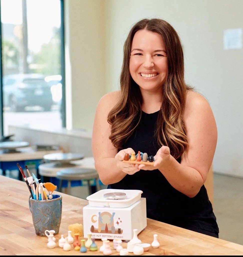 A smiling woman with long brown hair holding a tray of ceramic paint colors in a pottery studio.
