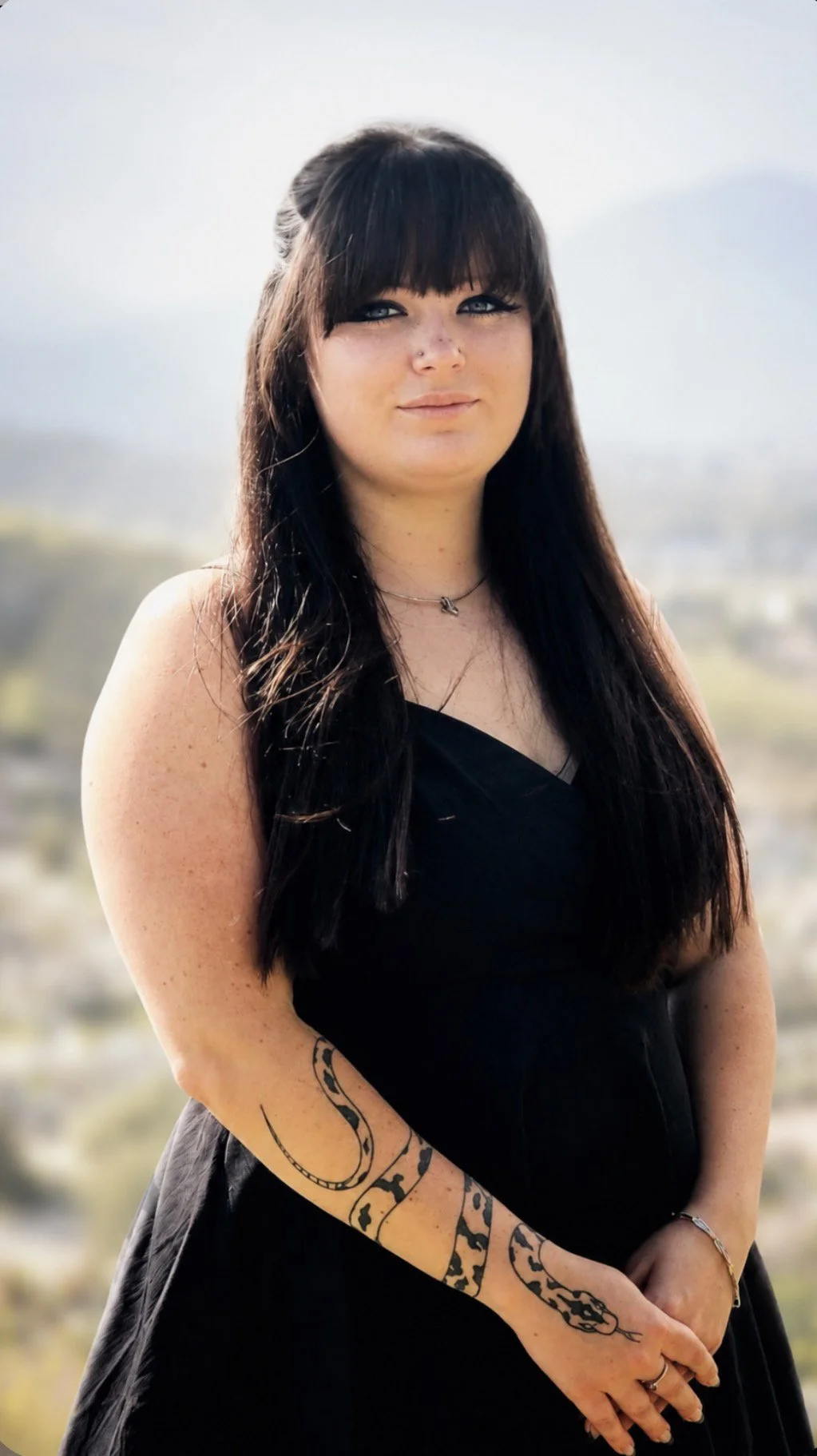 A young woman with long dark hair and bangs, wearing a black dress, standing outdoors with a blurred landscape background. She has tattoos on her left arm and a small necklace around her neck.