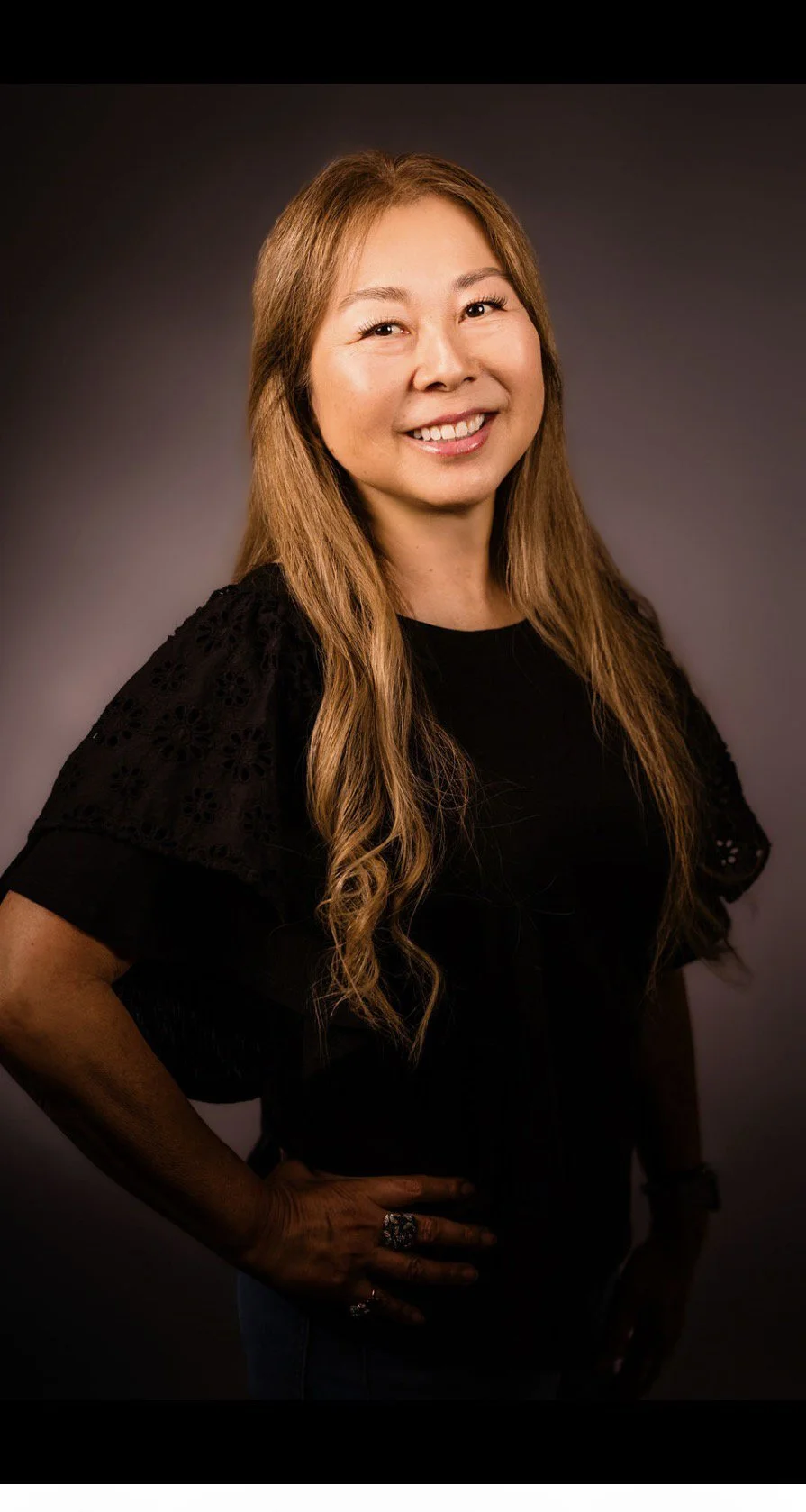 Smile woman with long blonde hair wearing a black top with lace details on sleeves posing against a dark background.