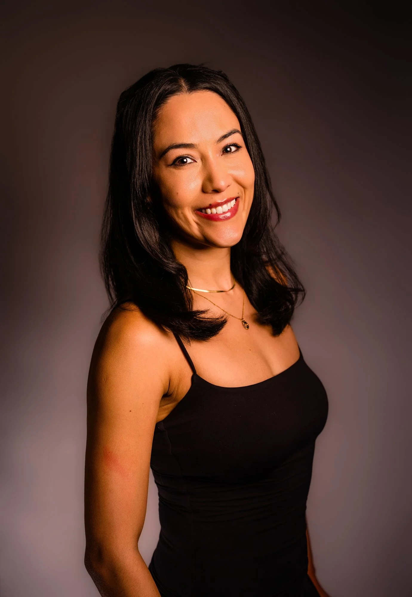 A woman with shoulder-length black hair wearing a black tank top and gold jewelry smiling at the camera against a dark gray background.