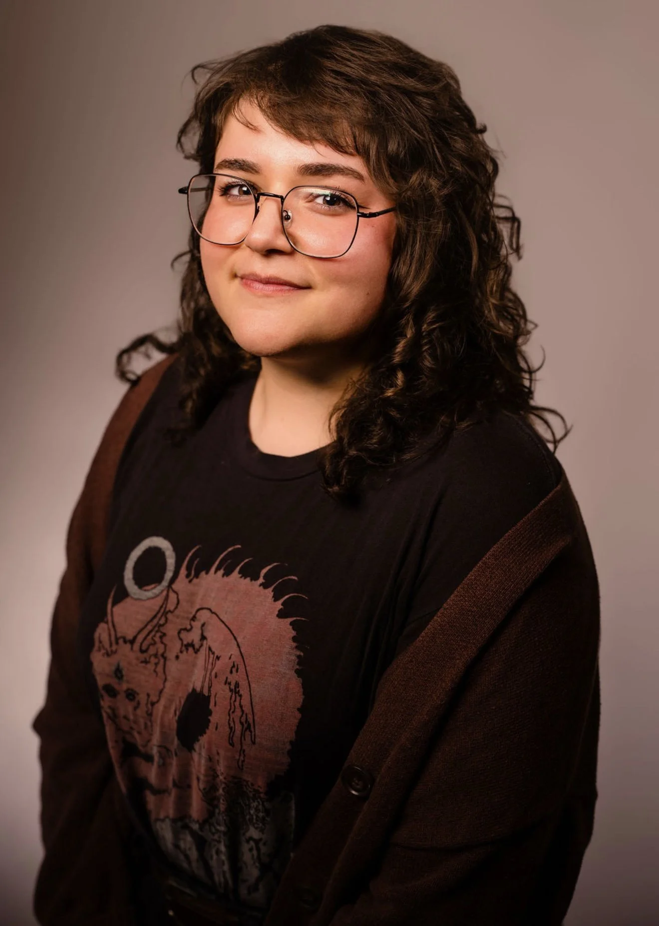 Portrait of a young woman with curly brown hair wearing glasses, a black graphic T-shirt with a skull design, and a brown jacket against a neutral background.