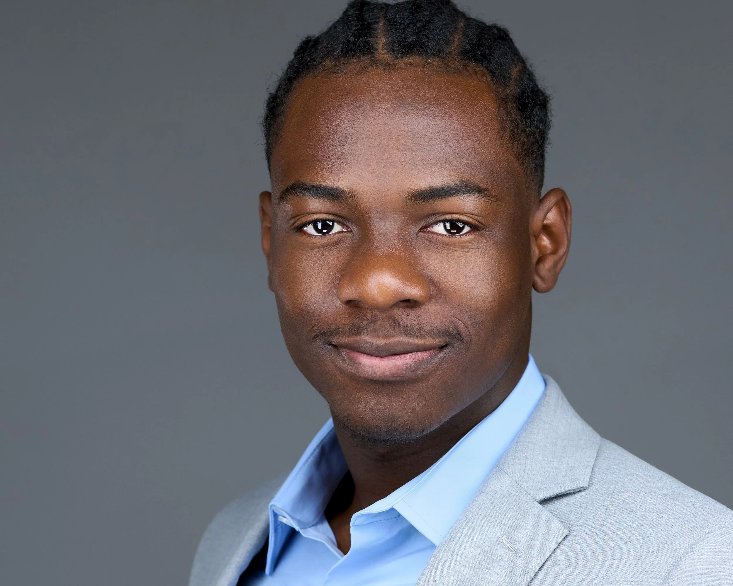 Headshot of a young man with dark skin, wearing a light gray suit and blue shirt, smiling slightly, with a gray background.
