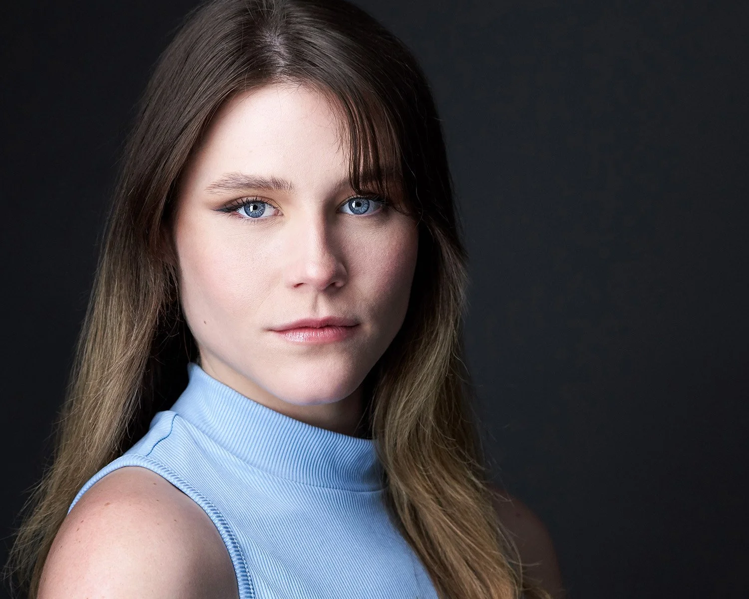 Close-up portrait of a woman with blue eyes, brown hair, and fair skin, wearing a light blue sleeveless top, against a black background.