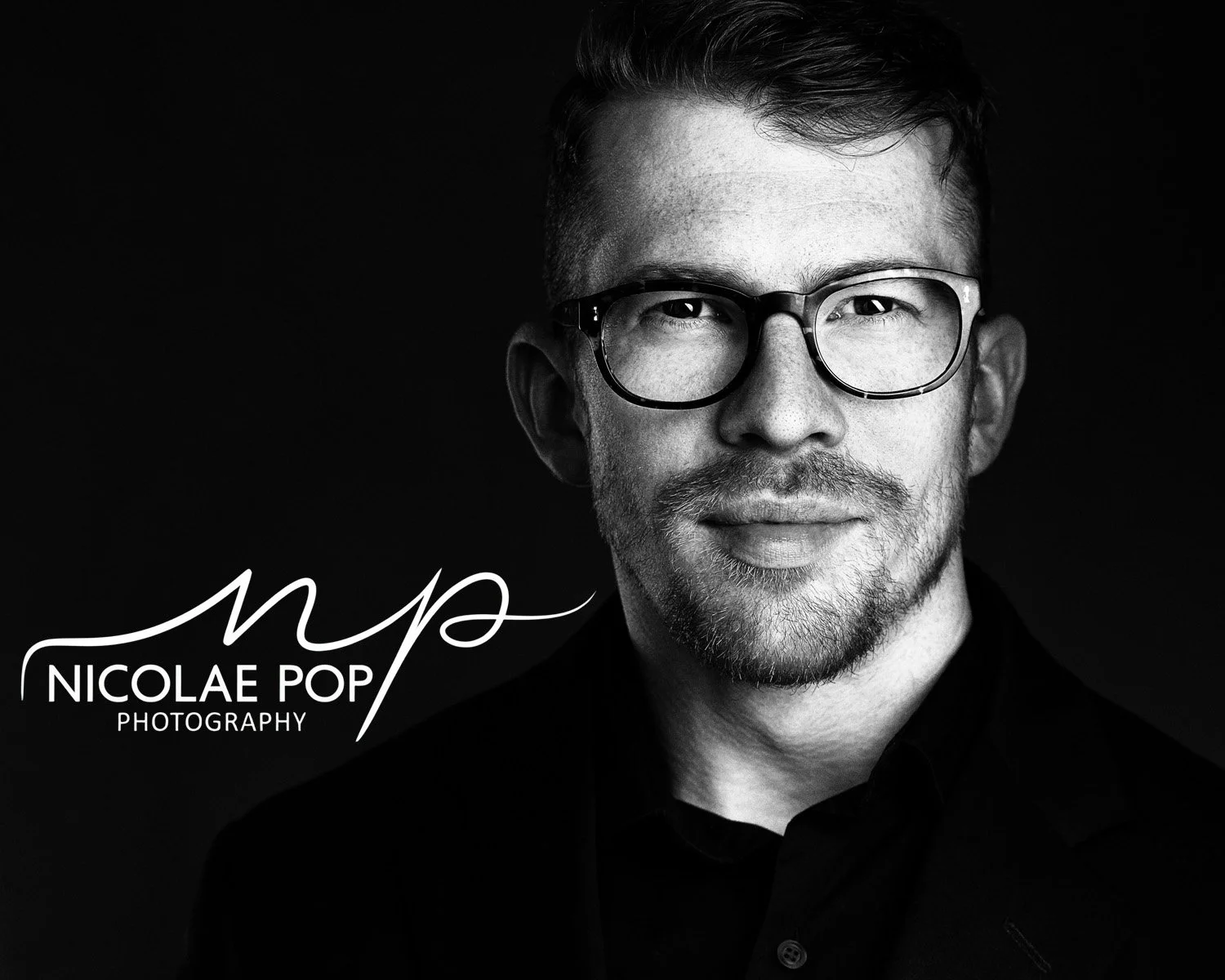 Black and white portrait of a man with glasses, short hair, and facial hair, wearing a dark shirt, against a dark background. The logo and name "Niclae Pop Photography" are in the lower left corner.