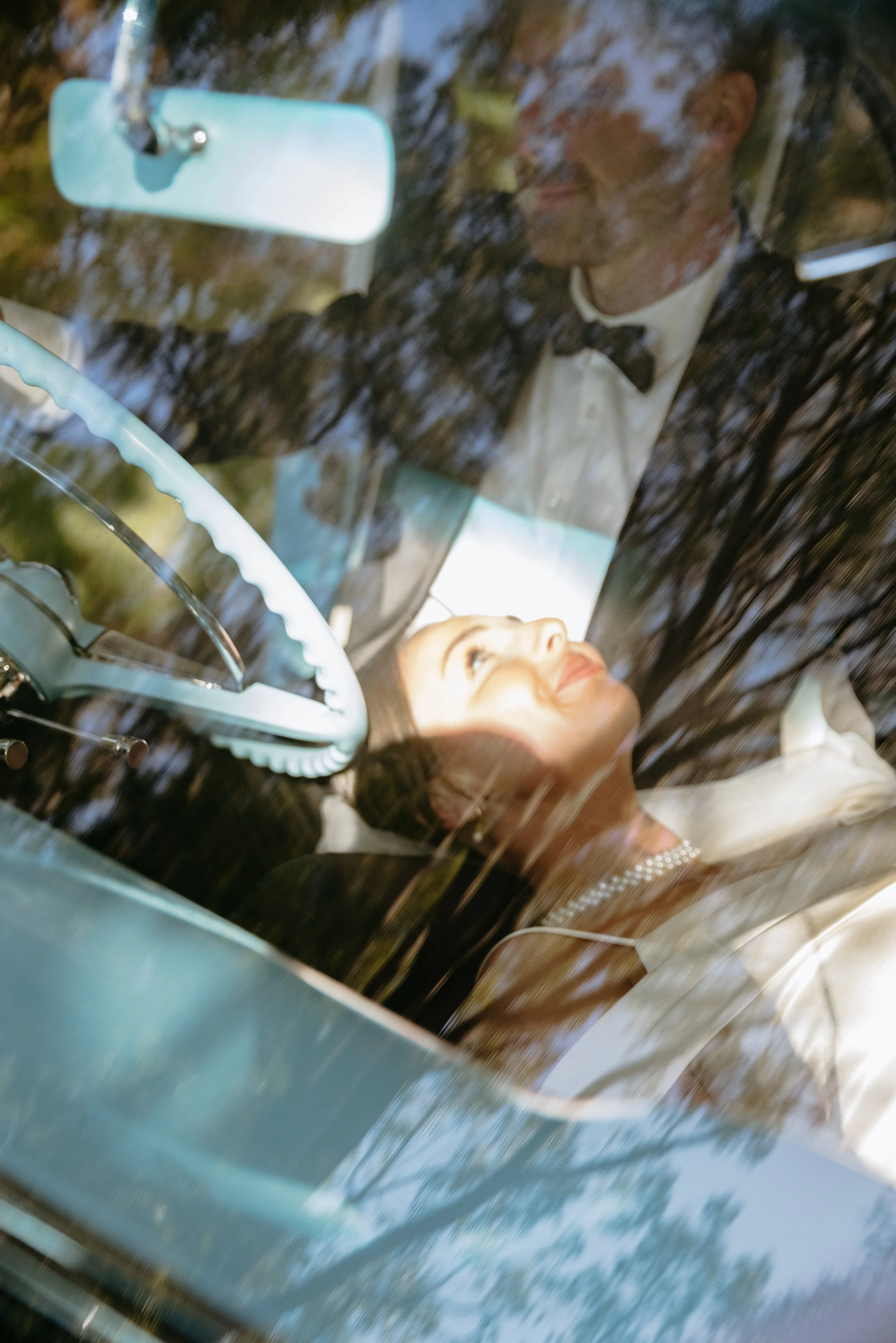 Bride and groom sitting inside a classic car, partially obscured by reflections on the windshield, with trees visible outside.