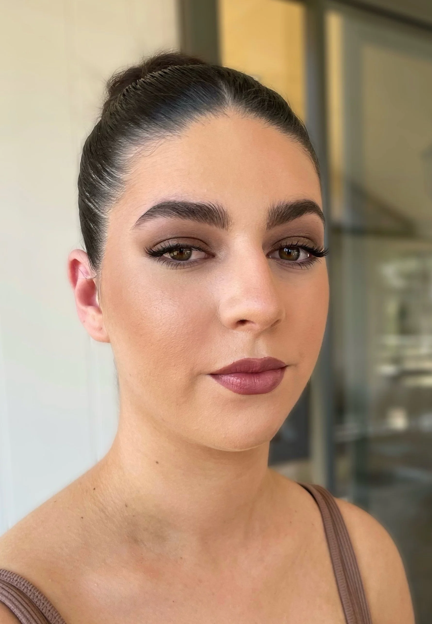 Close-up of a person with dark hair in a bun, wearing makeup and a neutral-colored top, indoors.