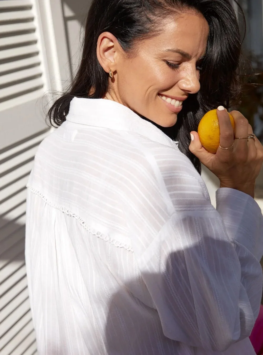 Woman in a white shirt holding a lemon, smiling, with sunlight creating shadows.