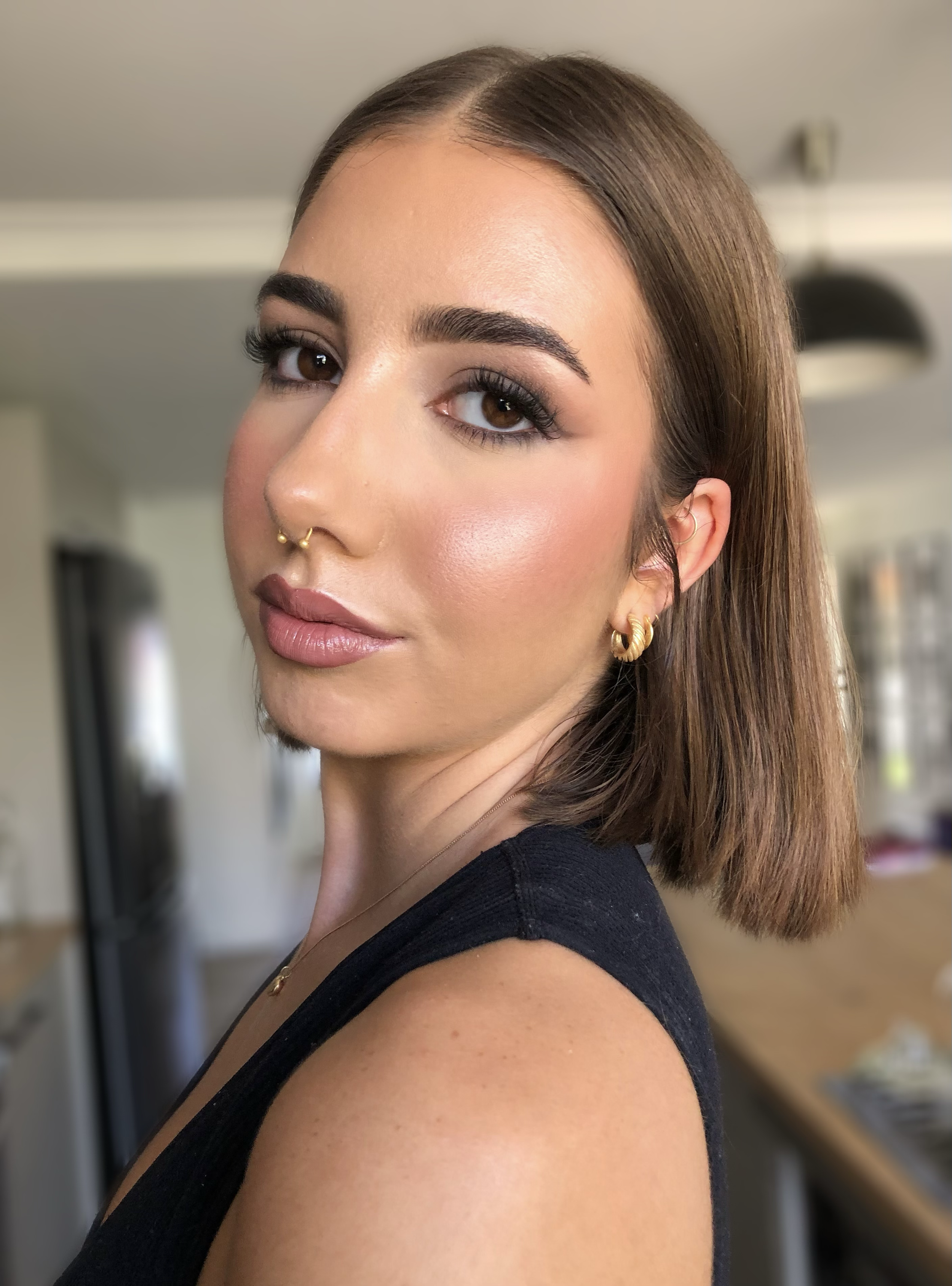 Person with short brown hair, wearing makeup, gold hoop earrings, and a nose ring, facing sideways in a modern kitchen.