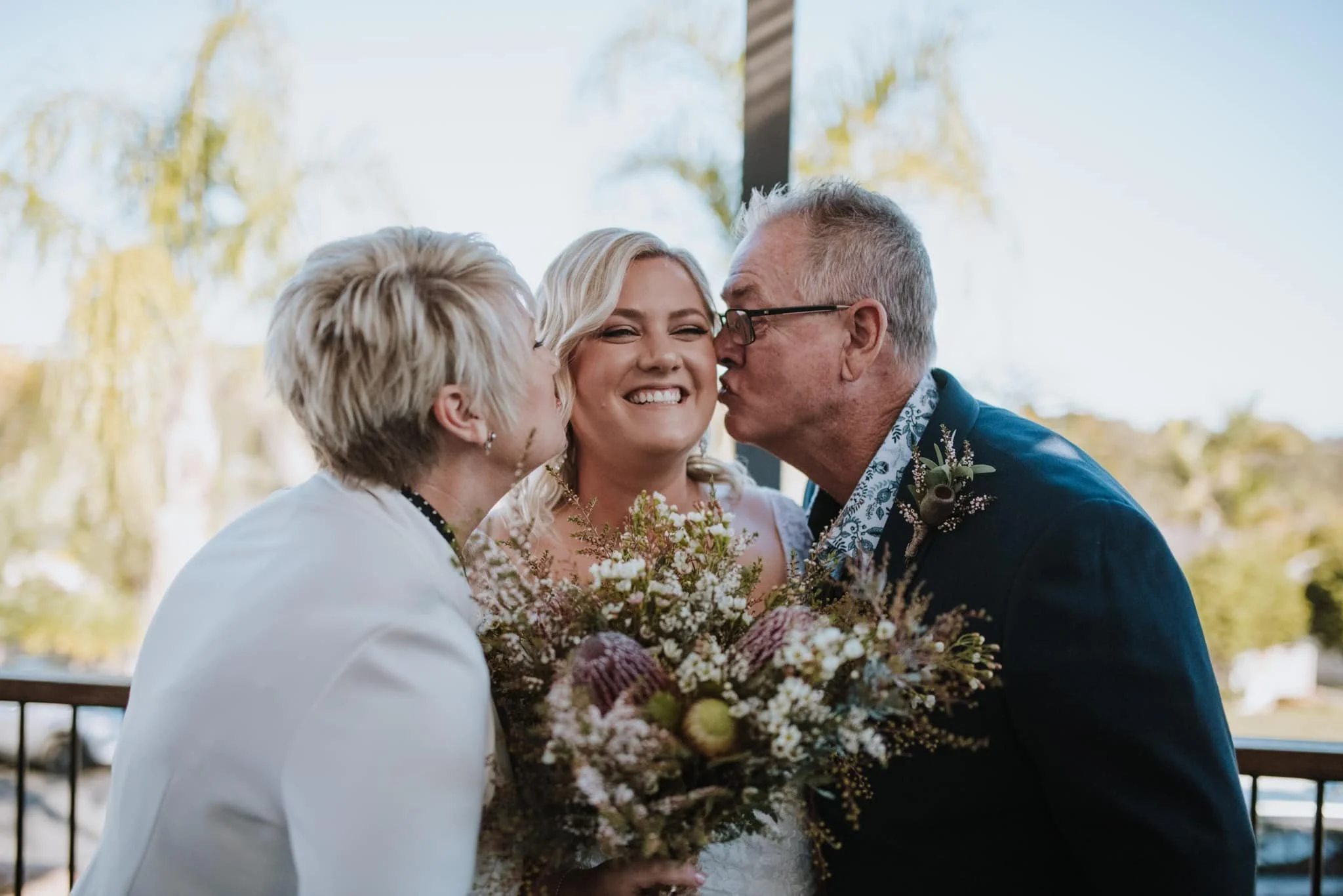 Bride smiling with floral bouquet, kissed on both cheeks by older couple outdoors.