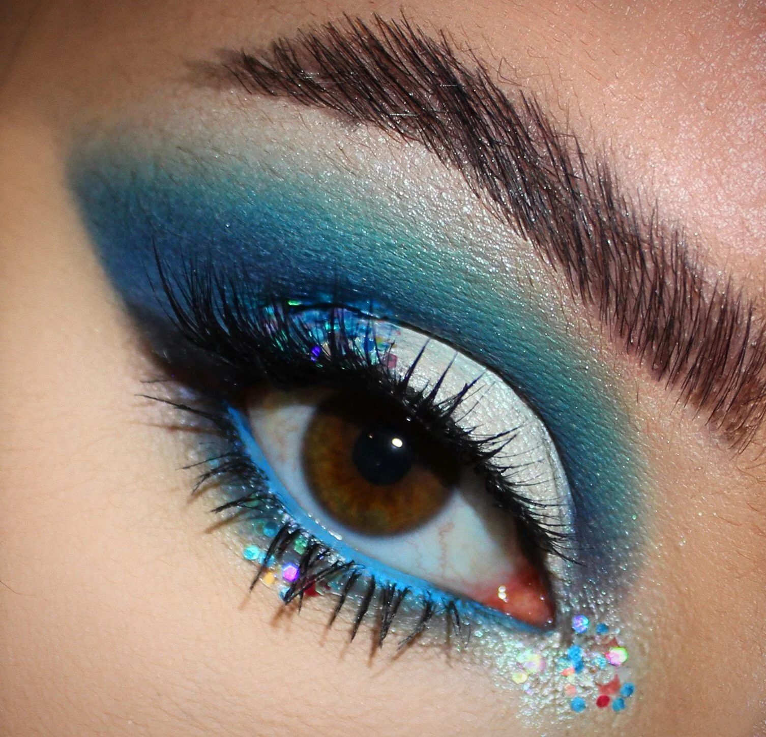 Eye with blue and white eyeshadow, thick black lashes, and colorful glitter accents.