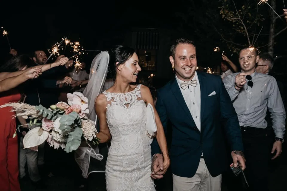 Bride and groom smiling surrounded by guests with sparklers at nighttime wedding celebration.