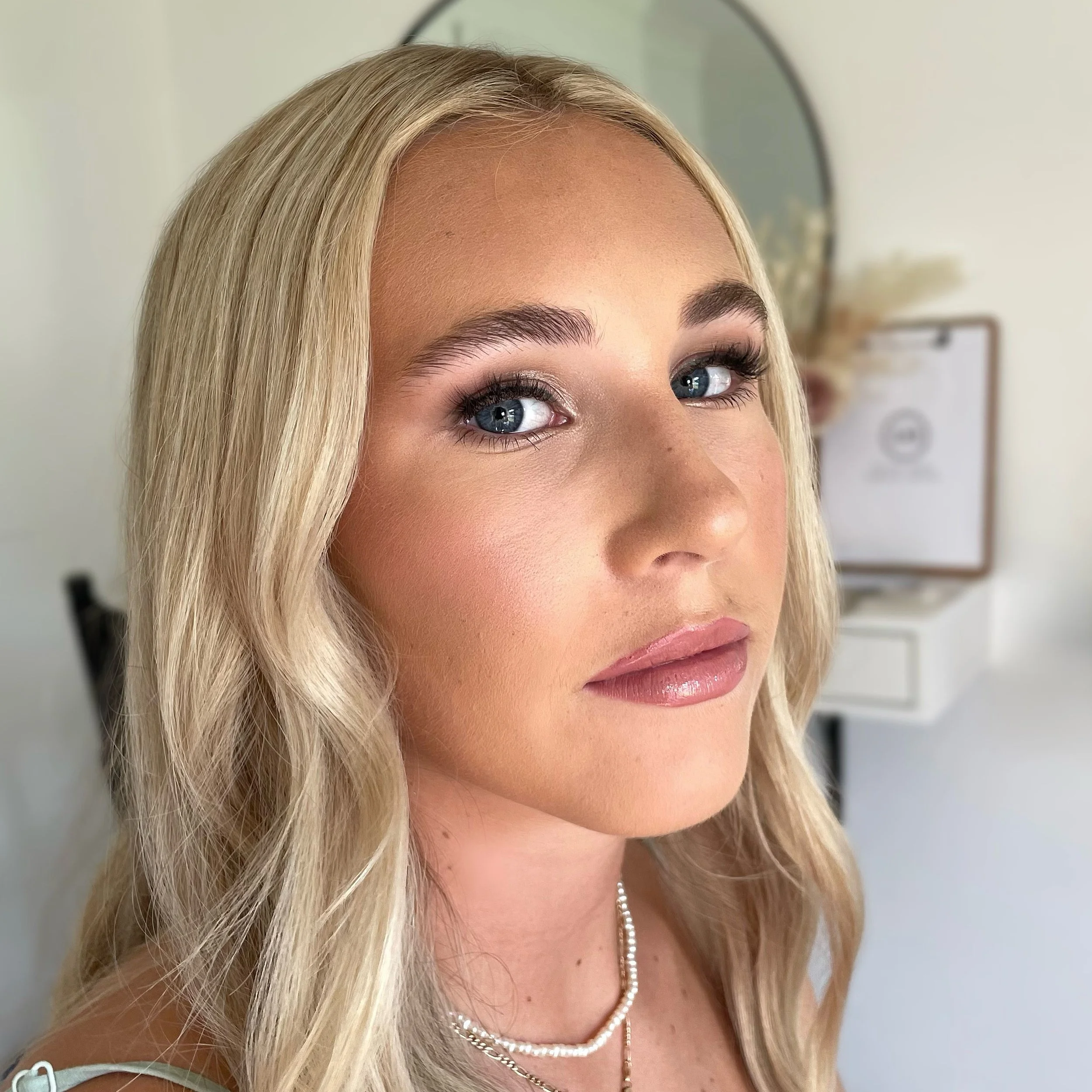 A woman with long blonde hair and makeup, wearing a necklace, looking towards the camera, with a mirror and blurred background.