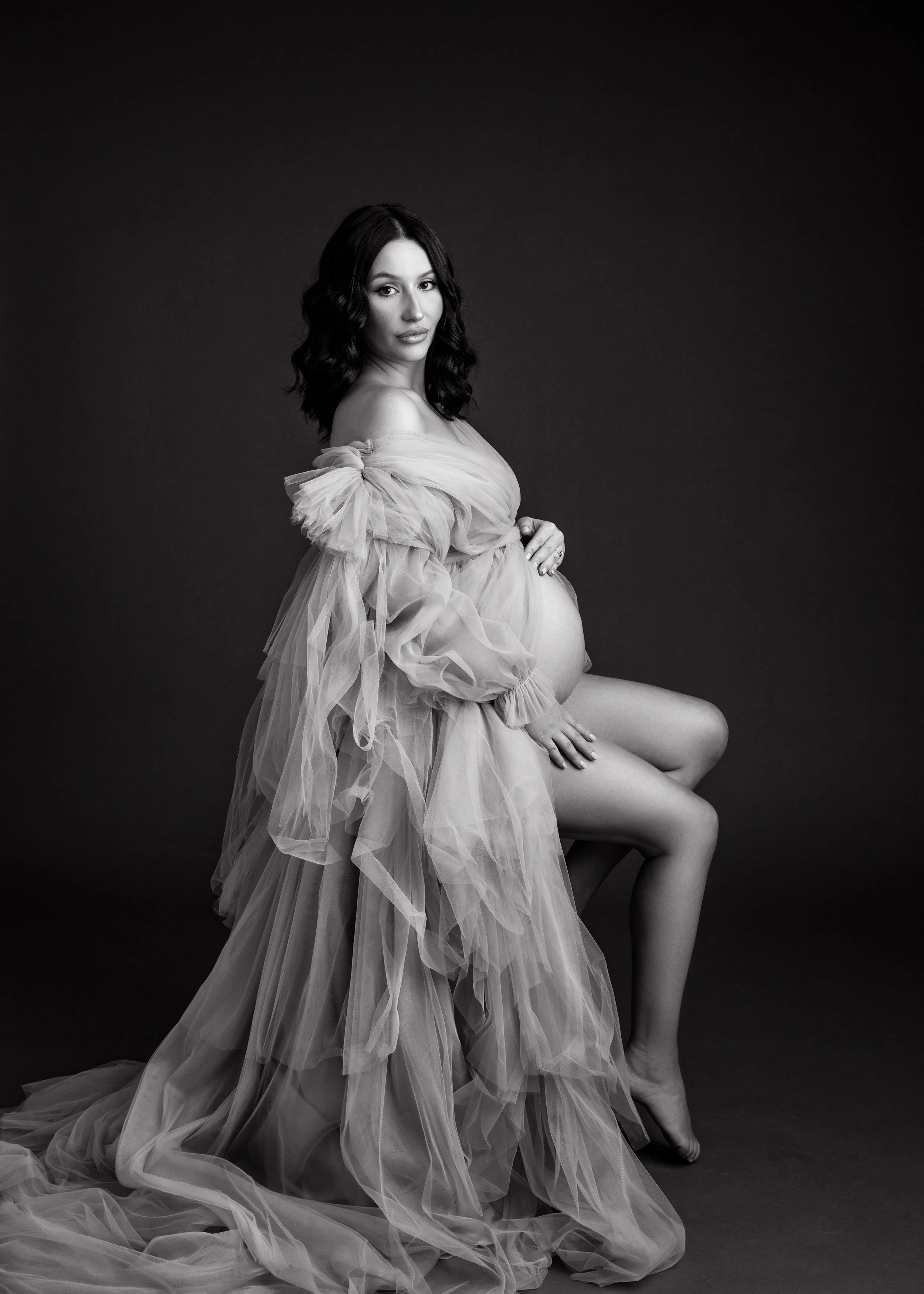 Black and white maternity portrait of a pregnant woman sitting on a stool, draped in a flowing, sheer gown, with one hand on her belly and the other resting on her knee.