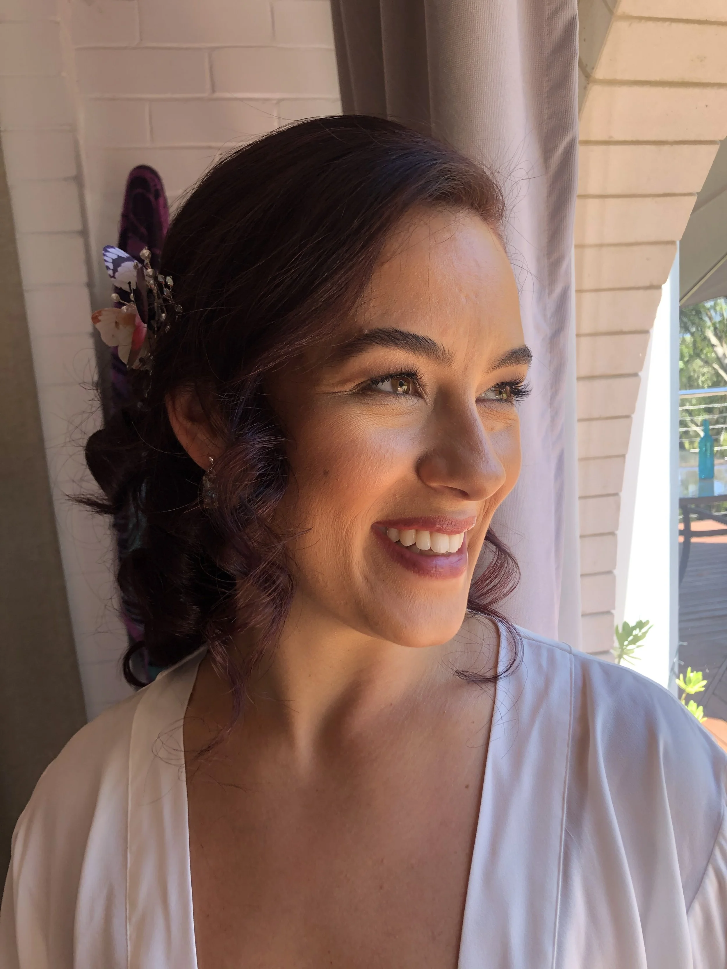 Smiling woman with curled hair and floral hair accessory, wearing a white robe, looking out a window.