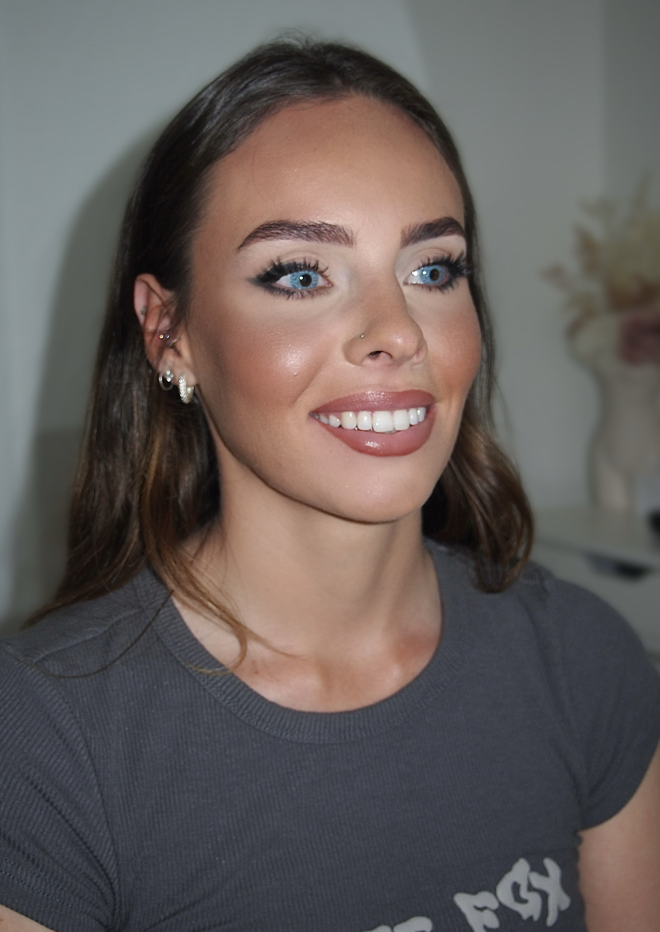 Smiling person with makeup, wearing earrings and a nose ring, in a gray shirt.