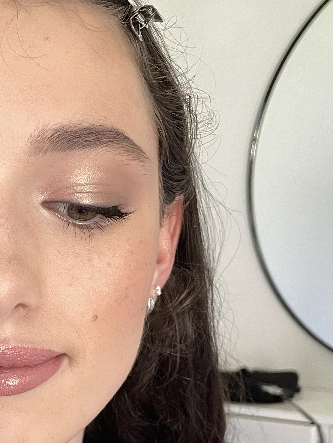 Close-up of a woman's face with polished makeup, showing her eye, eyebrow, and lips. She has a hair clip and wears earrings. A circular mirror is visible in the background.