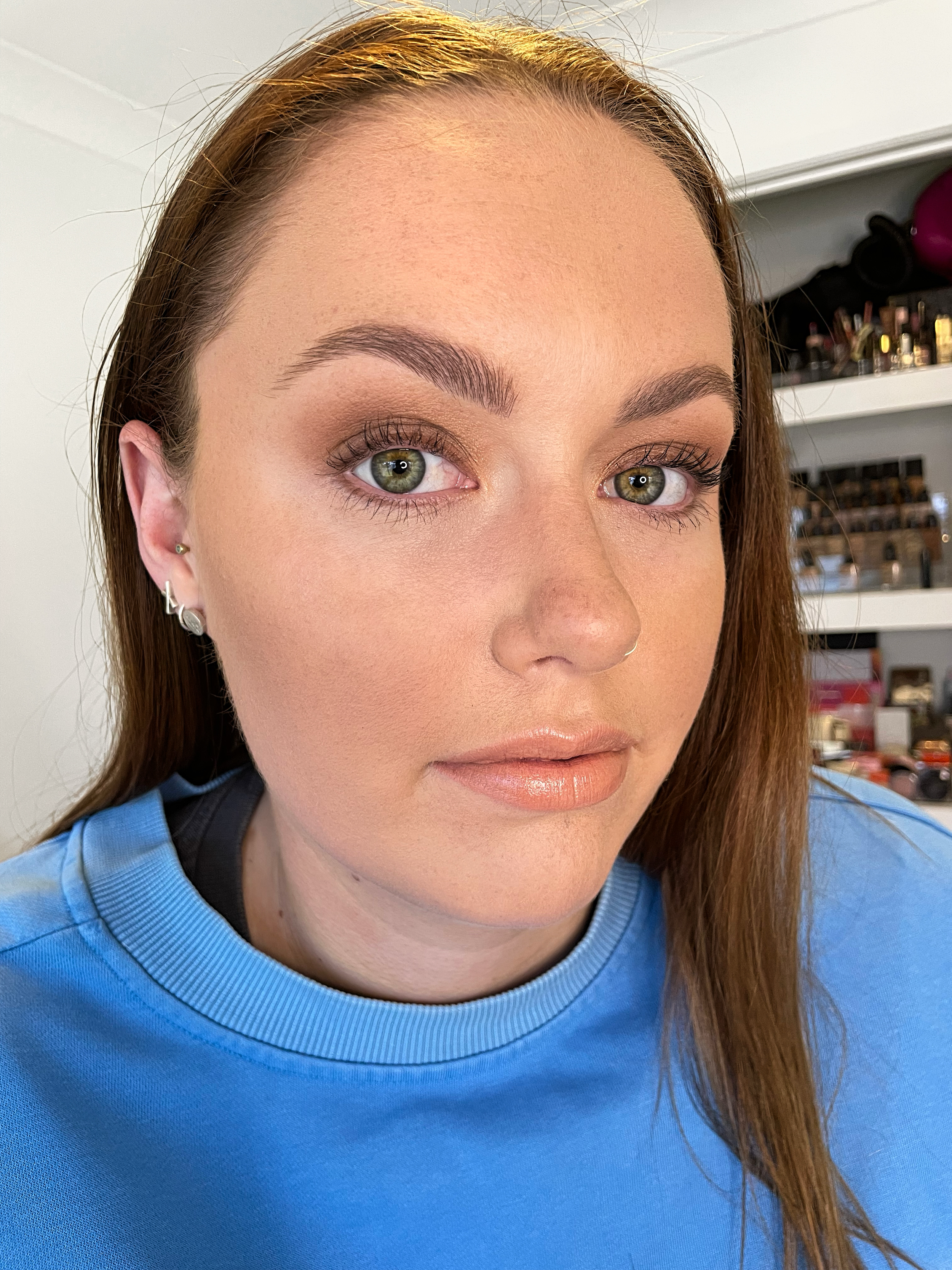 Person with light brown hair wearing a blue shirt, showcasing a makeup look with natural tones and glossy lips, indoors with makeup items in the background.