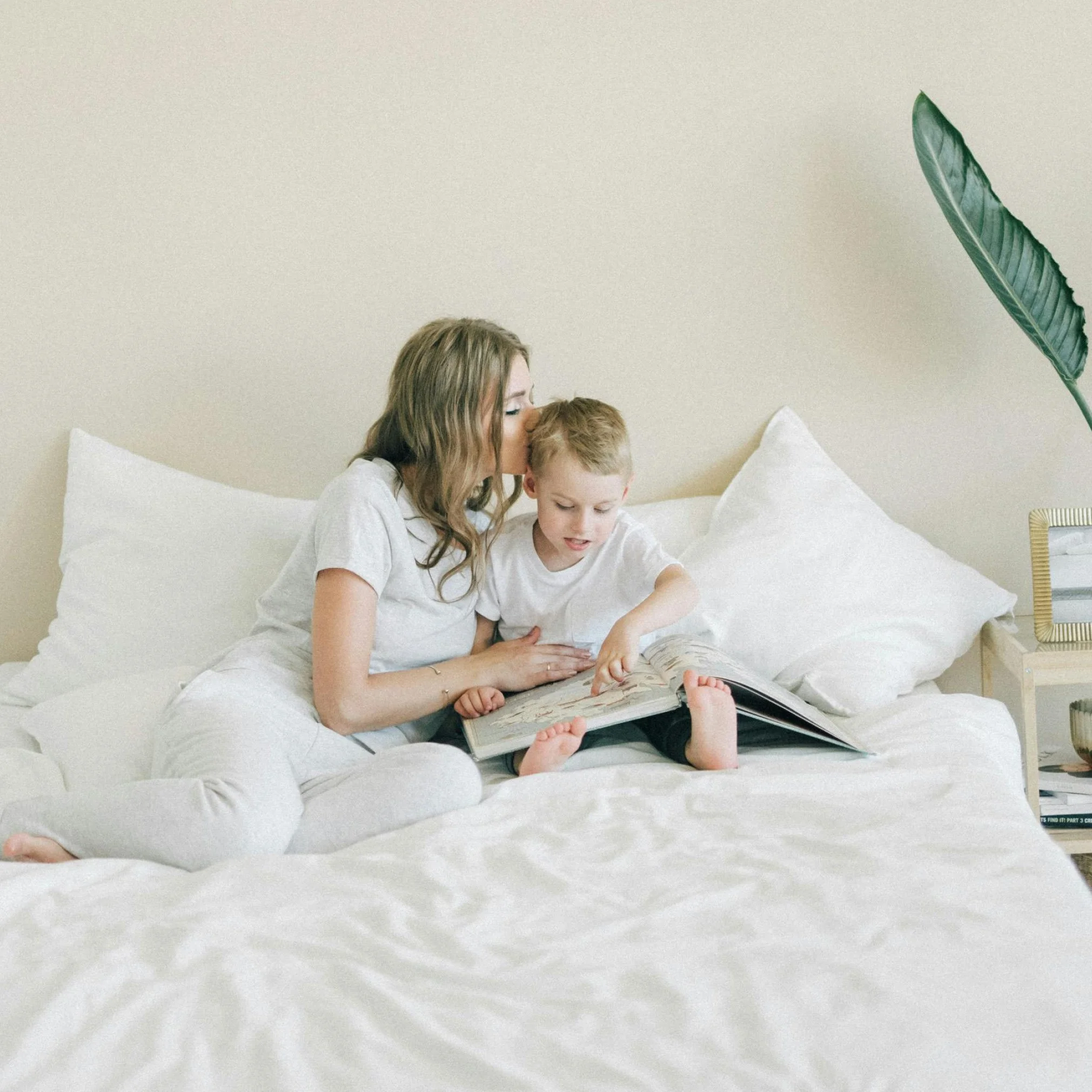 Mother wearing white sitting in bed with her young boy reading a book