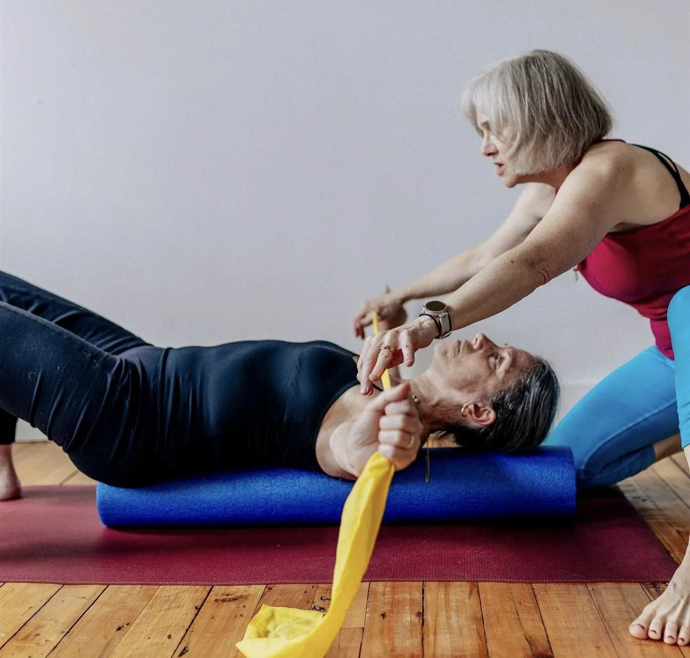 Two women are practicing yoga for the shoulders. One with osteoporosis is lying on her back on a foam roller, holding a yellow resistance band. The other is kneeling beside her, assisting with the stretch, both are engaged and focused.