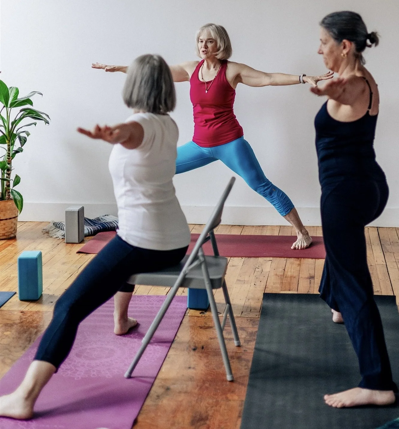 Yoga class 3 ladies with low bone density. One wearing a white t-shirt and black tights is seated in Warrior II pose, another in all black is standing in warrior II and the yoga teacher demonstrates warrior II wearing a fusia top and turquoise tights