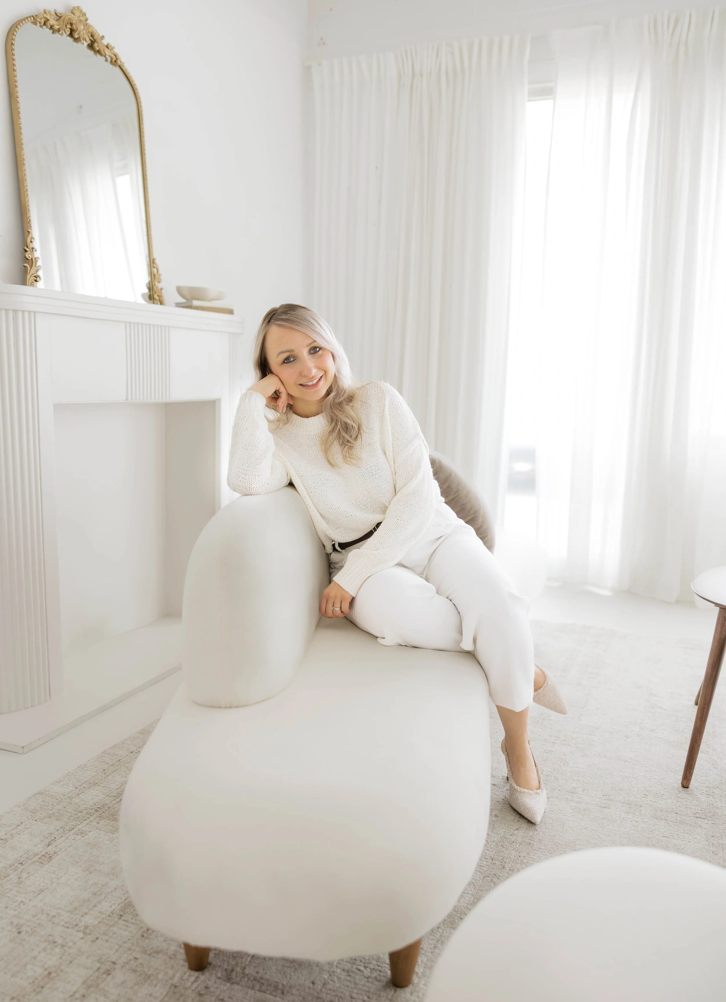 A woman sitting on a white modern chair in a bright, white room with white curtains, a white fireplace, and a large mirror with an ornate gold frame, smiling and resting her head on her hand.