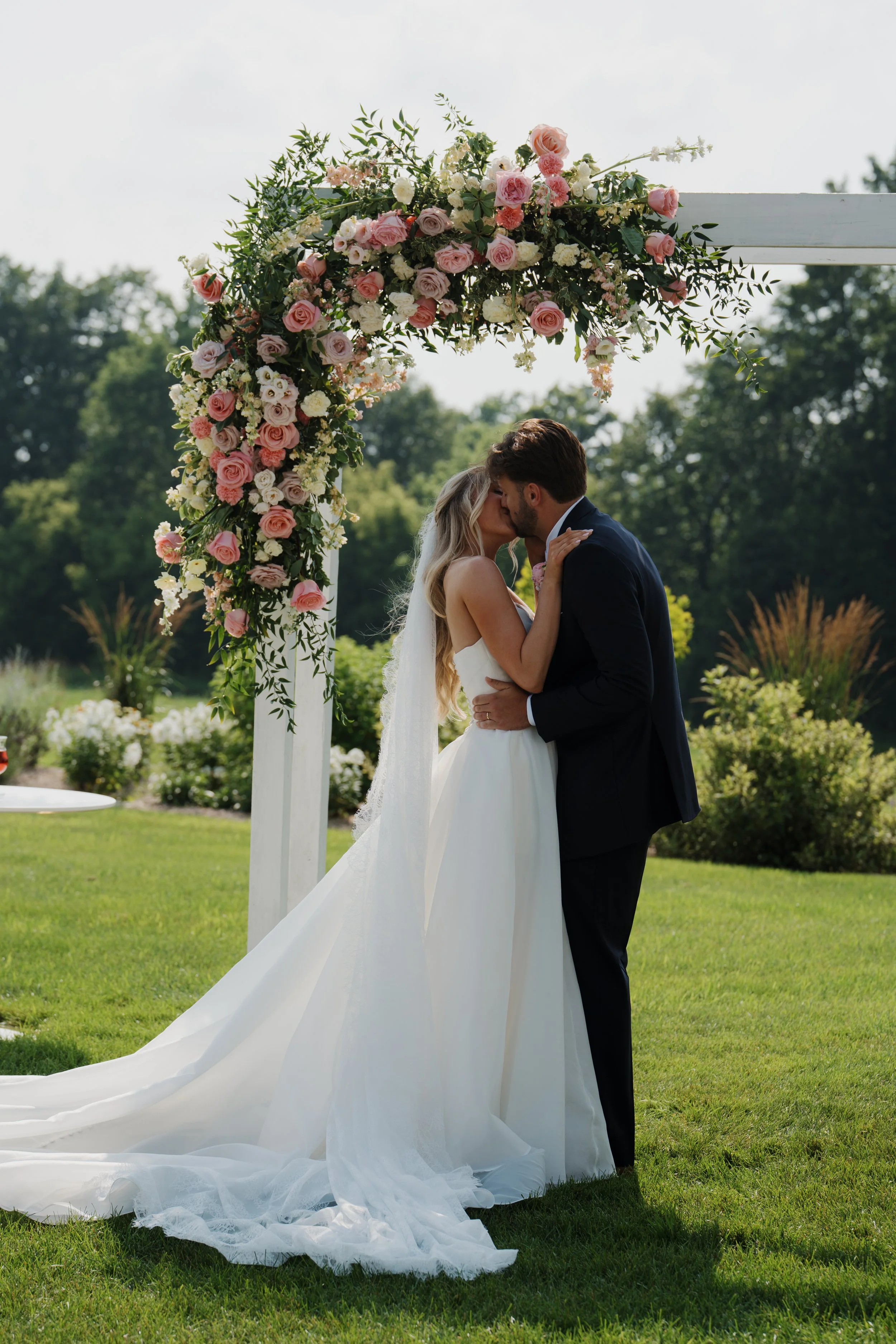 A bride and groom kissing under a floral wedding arch outdoors on a sunny day.